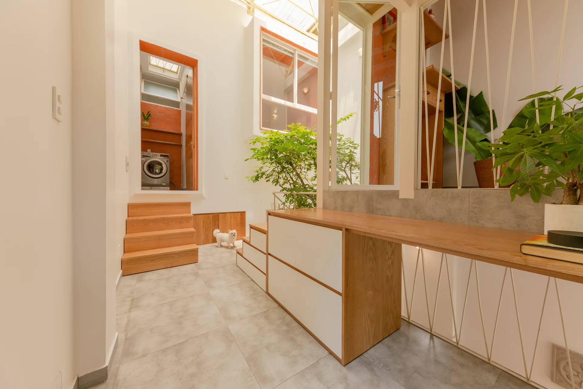 A modern split-level interior space with grey tile floors, featuring a built-in wooden desk, tiered wooden steps, and a view into a laundry area.