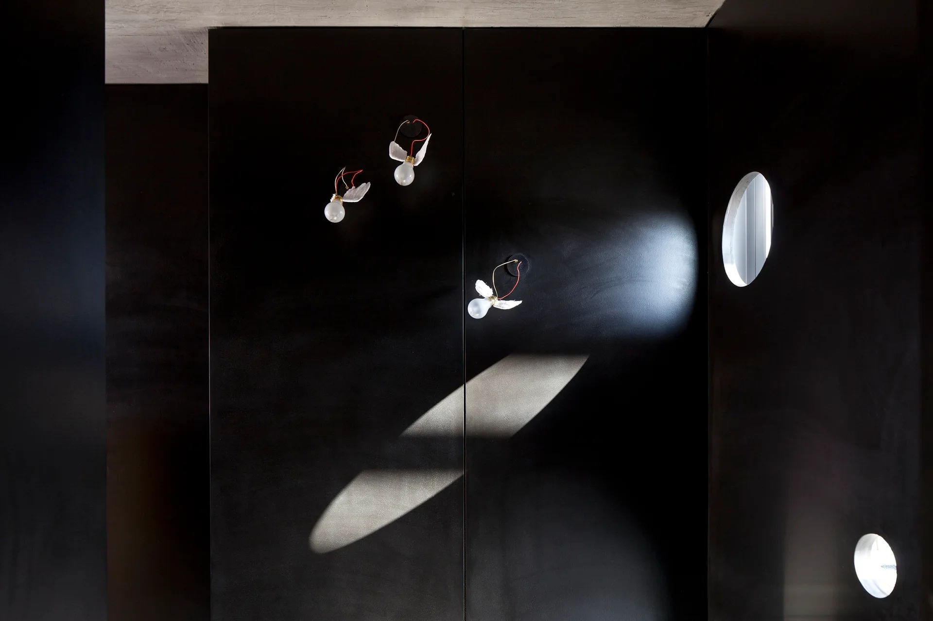 Three winged light bulb fixtures mounted on a matte black wall with small circular openings.