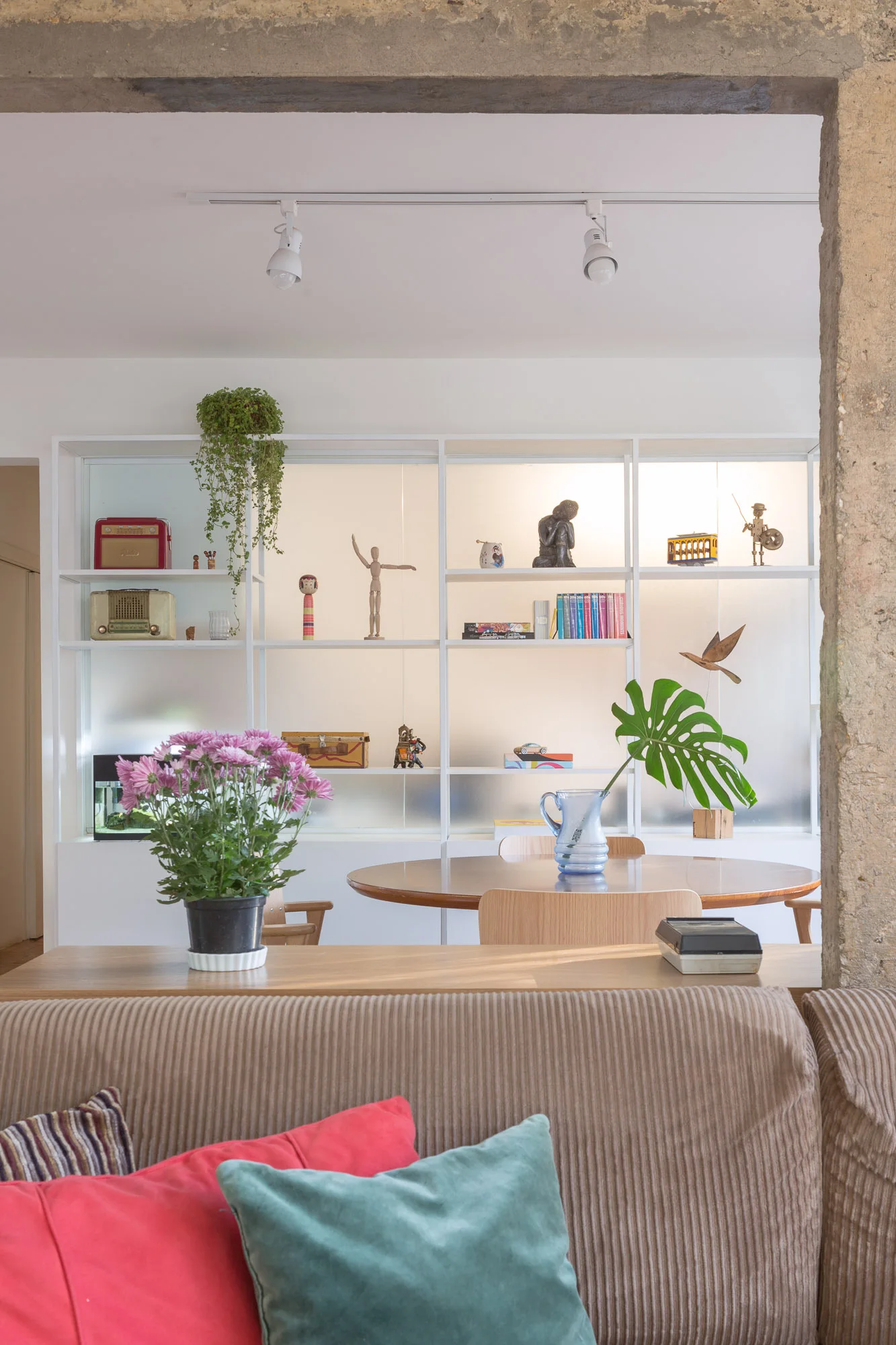 A dining room with a round wooden table, chairs, and a large white backlit shelving unit, viewed through a rough concrete archway.