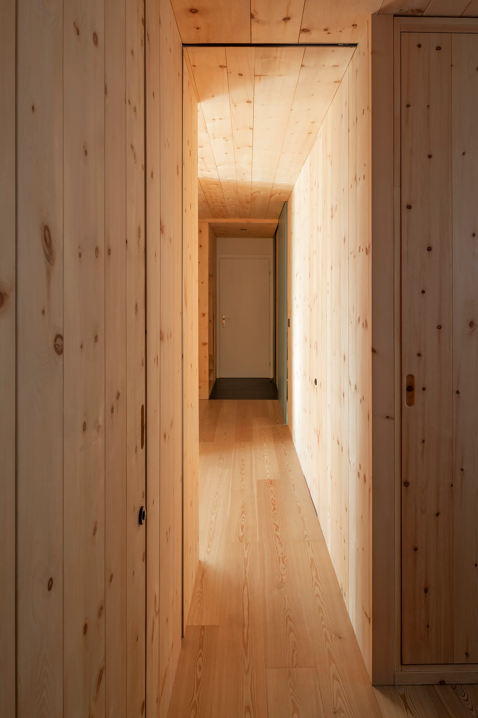 A narrow hallway featuring knotty pine wood paneling on the walls, ceiling, and floor with flush integrated doors and a white door at the far end.