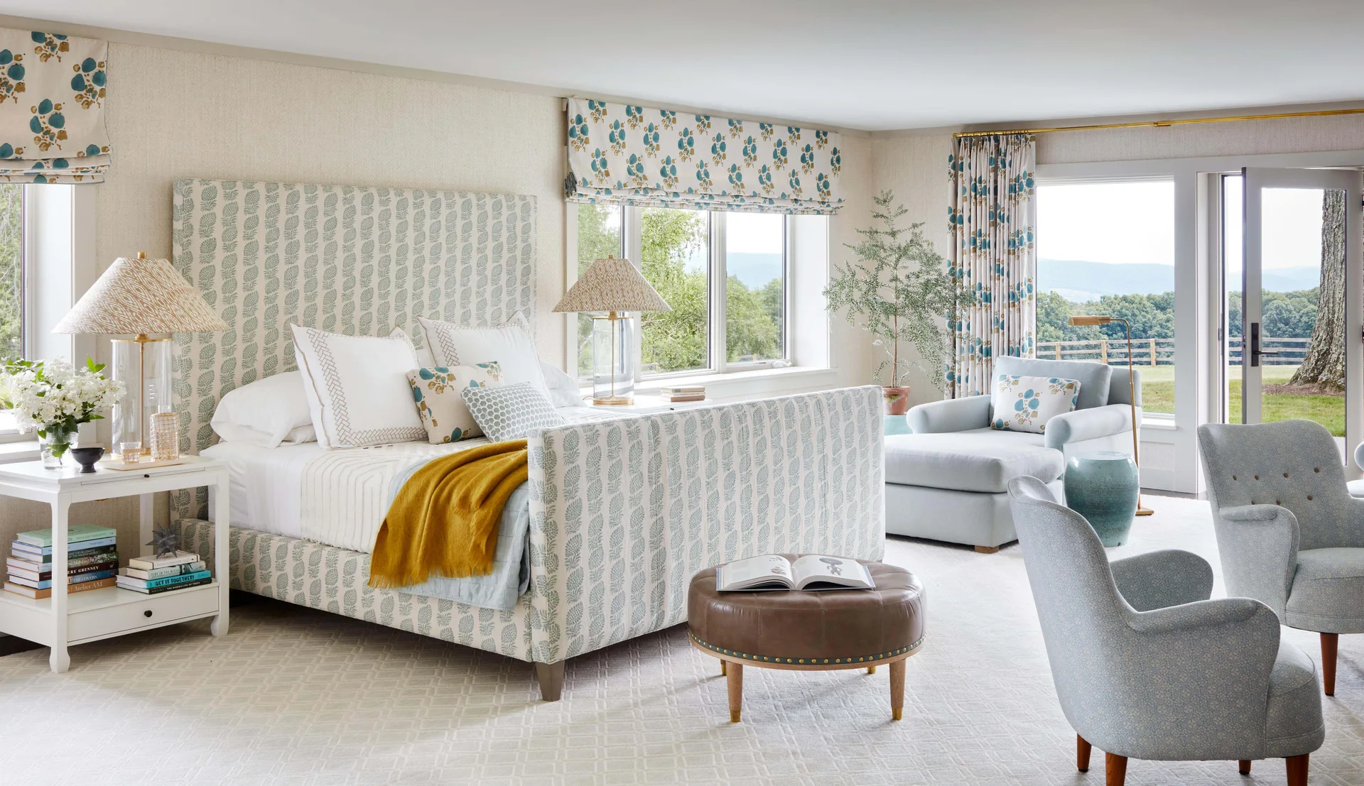 A light-filled bedroom features an upholstered patterned bed, matching window treatments, a light blue sitting area, and a round leather ottoman.
