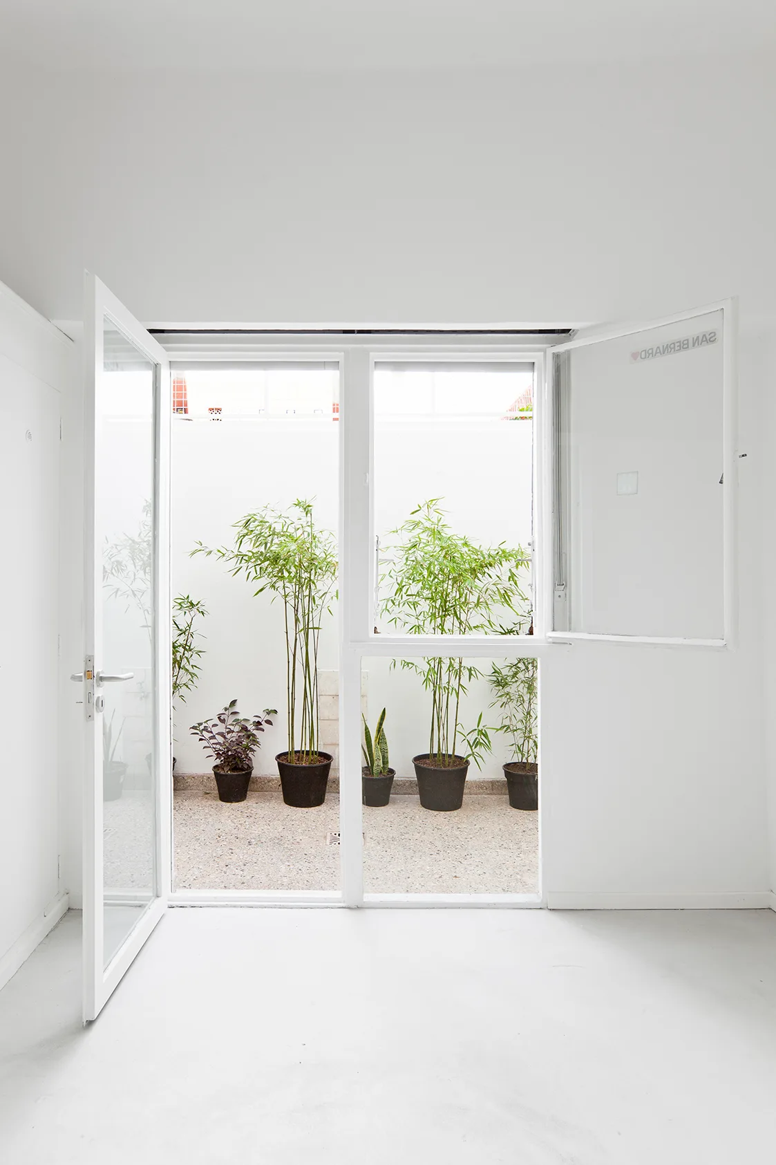 A bright white minimalist room features an open glass door looking out onto a small gravel courtyard with potted green bamboo plants.