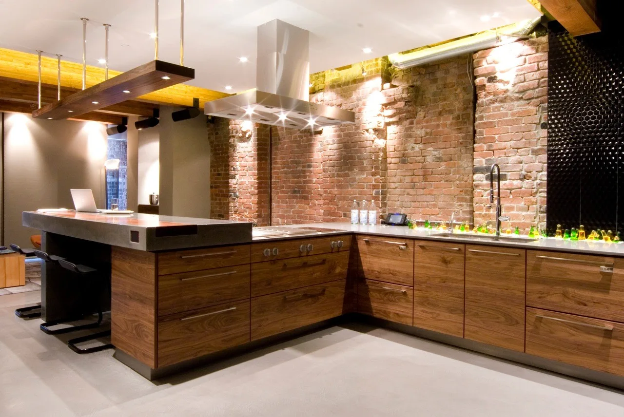 A modern kitchen features lower walnut cabinets, an exposed brick wall, a stainless steel range hood, and a concrete floor.