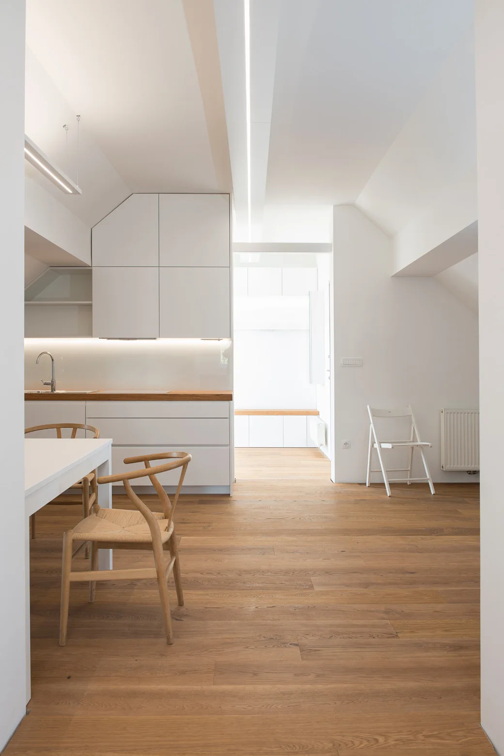A minimalist white kitchen features handleless cabinets, a natural wood countertop, and matching wooden wishbone chairs.