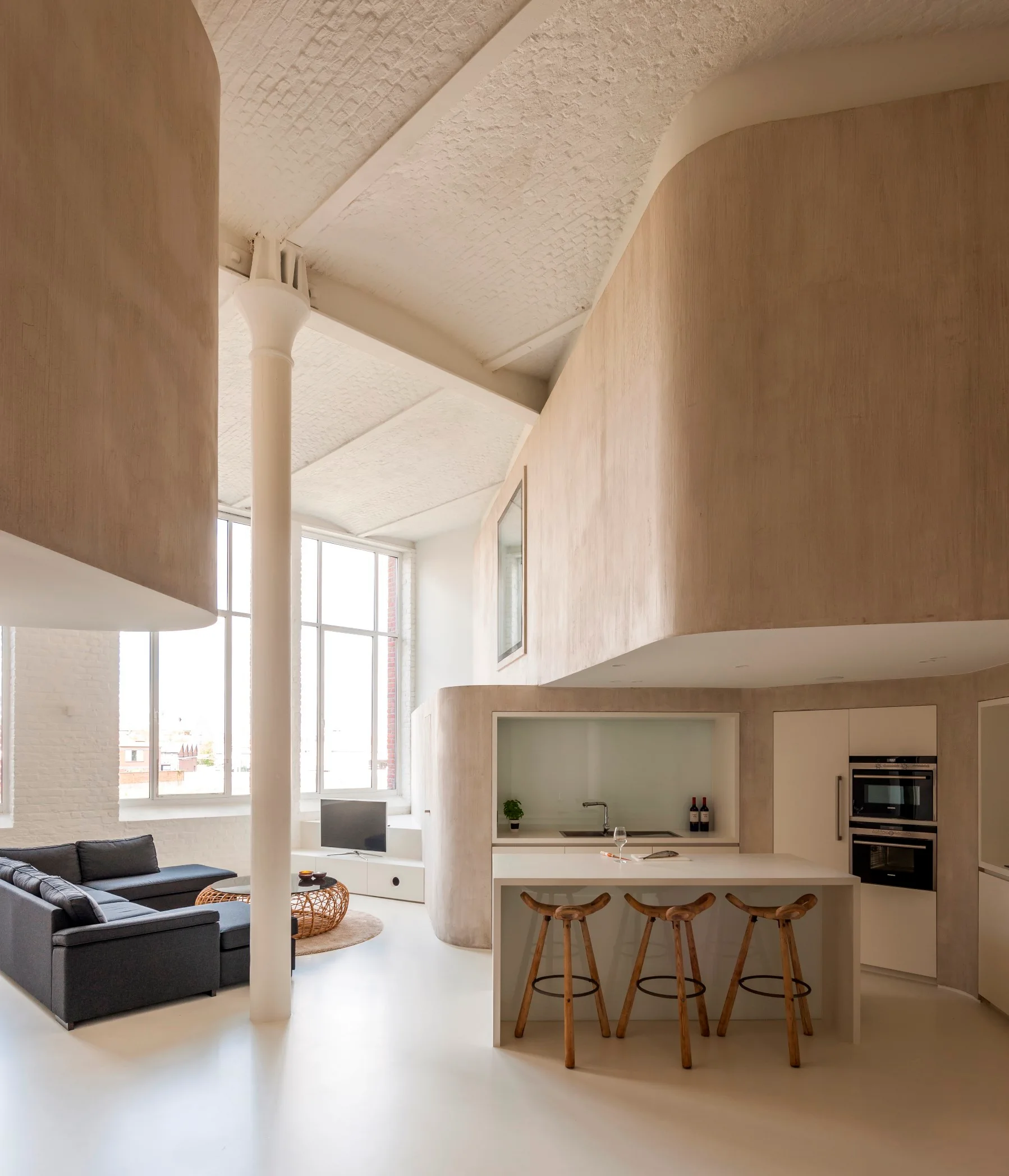 A minimalist loft space featuring curved plaster walls, a kitchen island with wooden stools, a white column, and a dark gray sectional sofa.