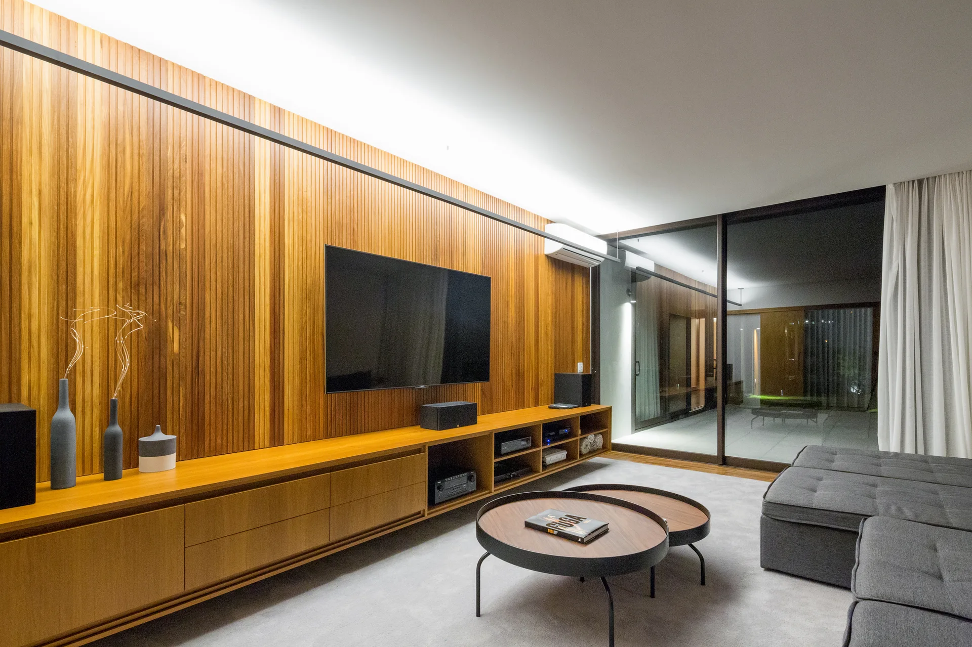 A modern living room features a vertical wood-slatted wall with a mounted television, a matching long low console, and a grey sectional sofa.