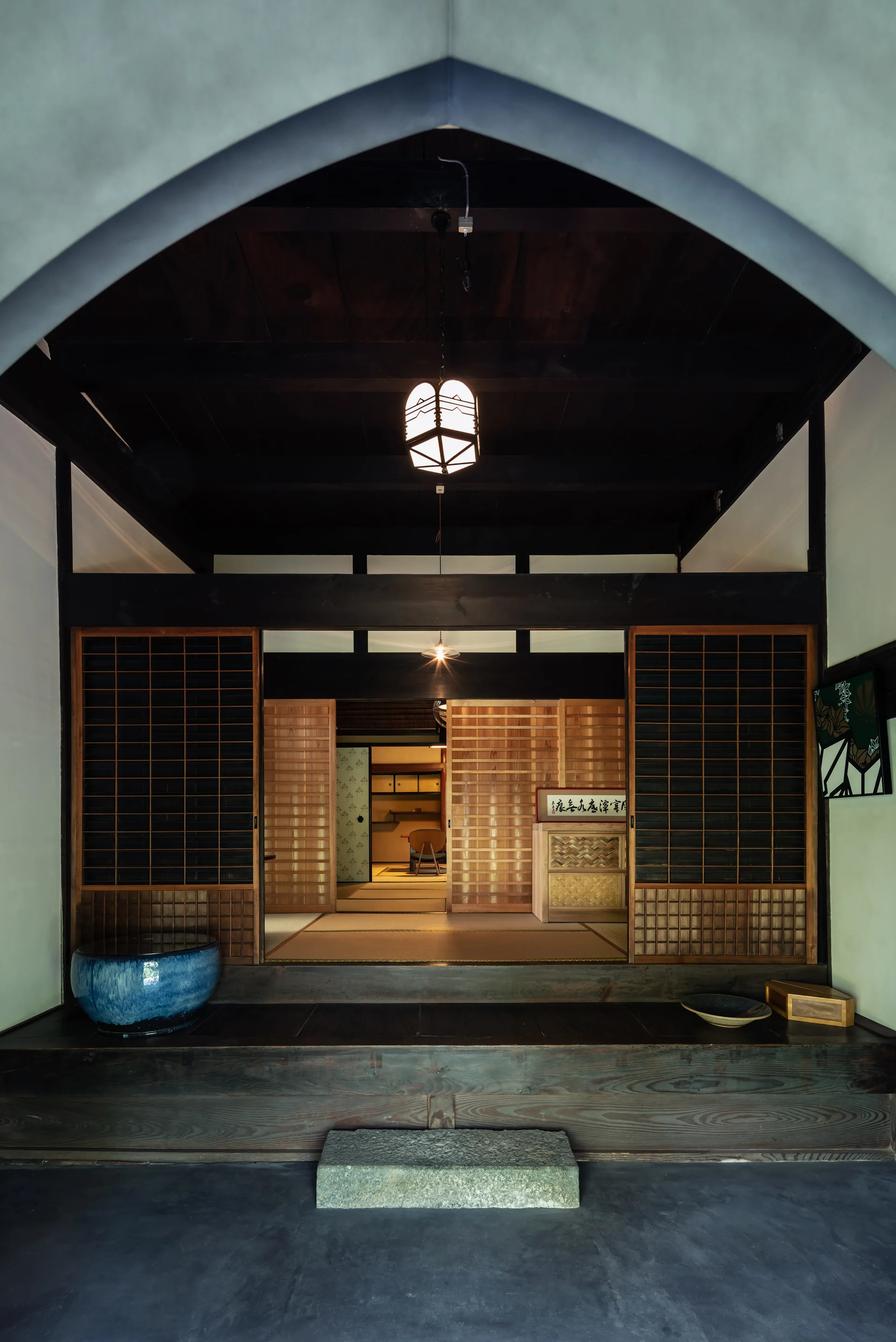 A traditional Japanese entryway framed by a pointed archway, featuring dark wood ceiling beams, sliding lattice doors, and a raised wooden step.