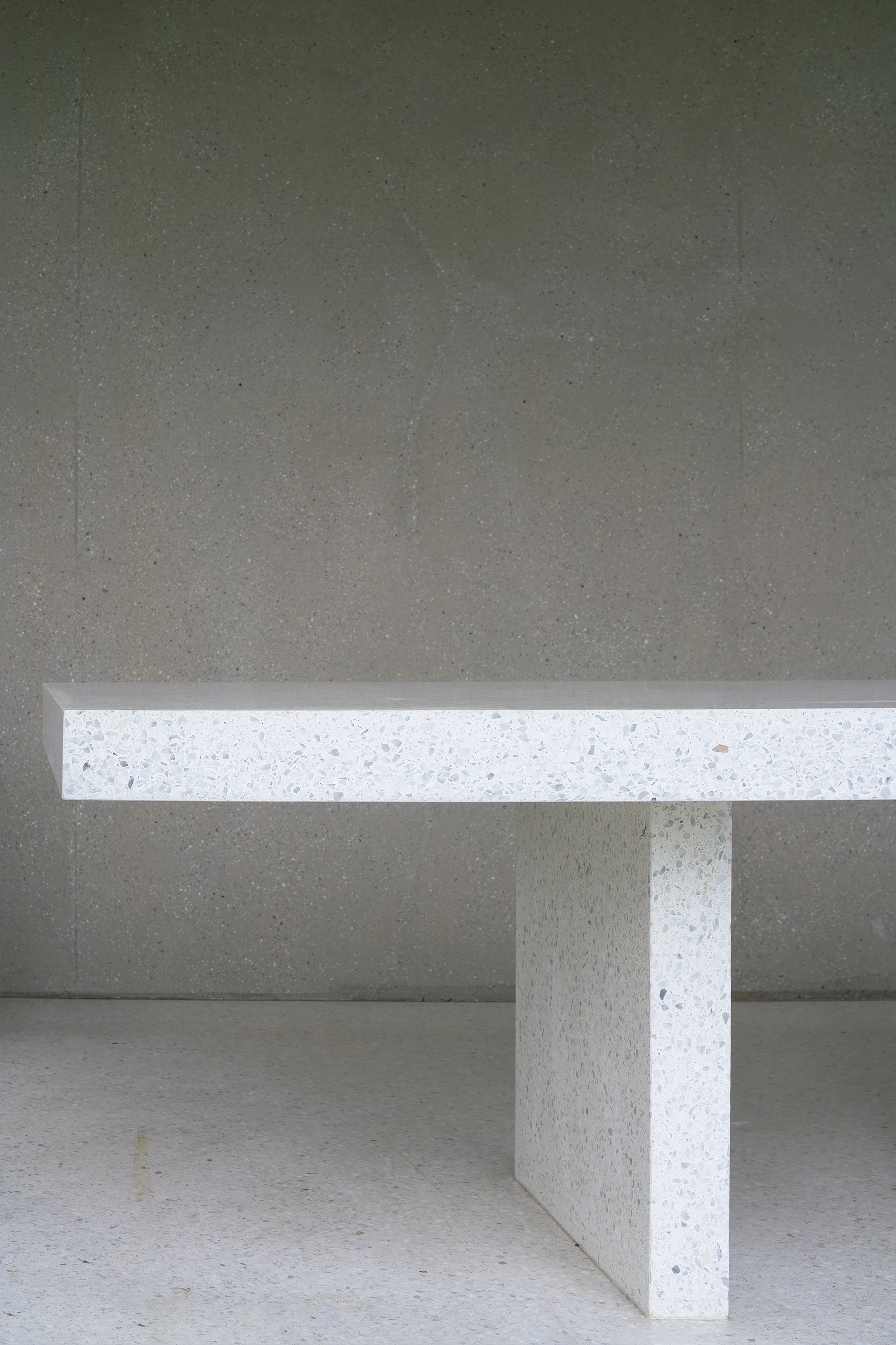 A modern white terrazzo table with a thick slab pedestal leg stands against a textured gray plaster wall on a light gray floor.
