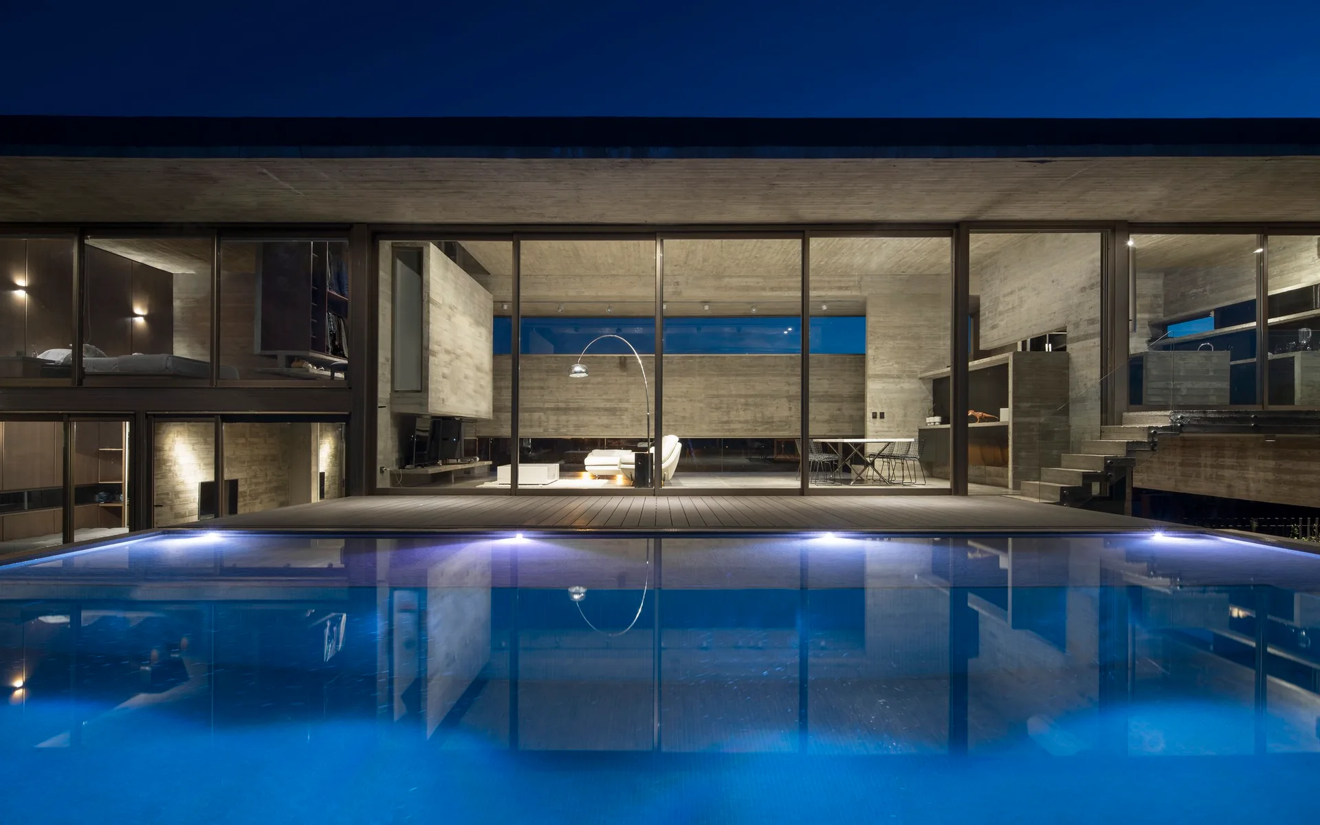 A minimalist modern concrete home at night featuring floor-to-ceiling glass walls that look out onto a glowing blue swimming pool.