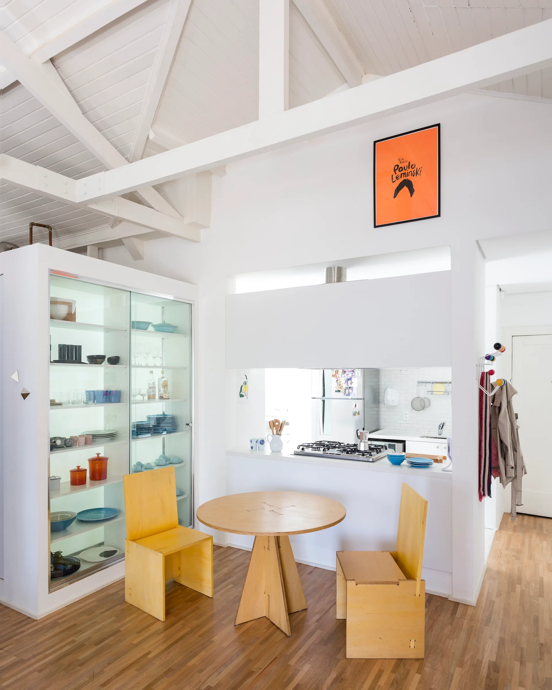A white kitchen and dining area features a circular plywood table with matching chairs next to a large glass-fronted display cabinet.