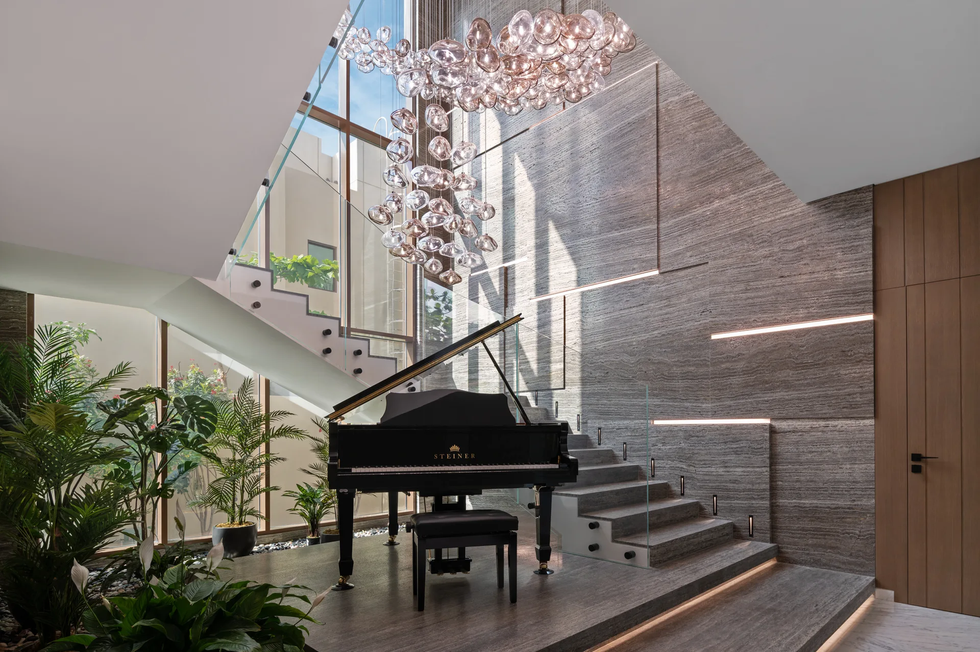 A black grand piano sits on a grey stone landing next to a staircase with glass railings and a large glass bubble chandelier hanging above.