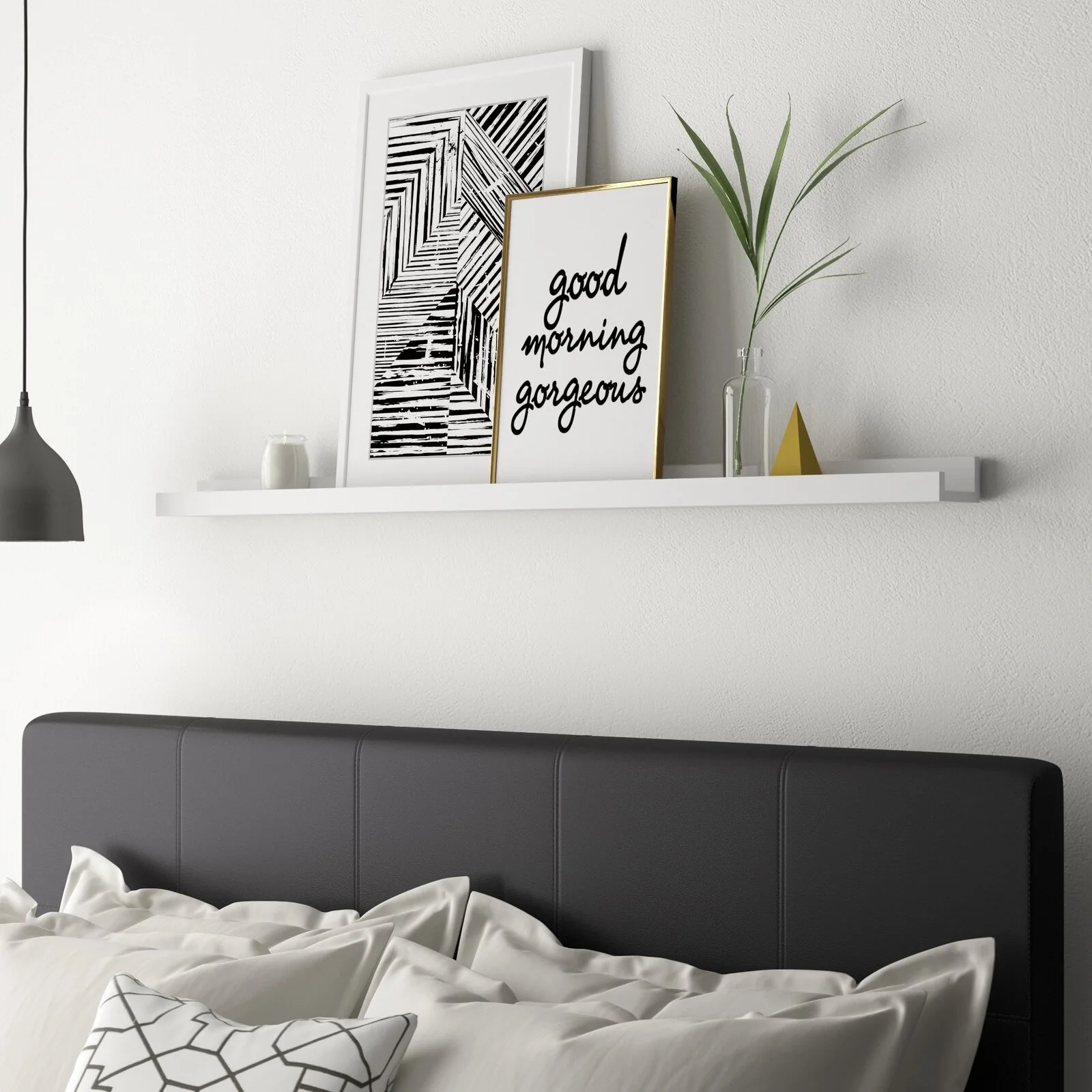 A minimalist bedroom vignette features a dark headboard below a white floating shelf holding two framed art prints and a small glass vase with greenery.