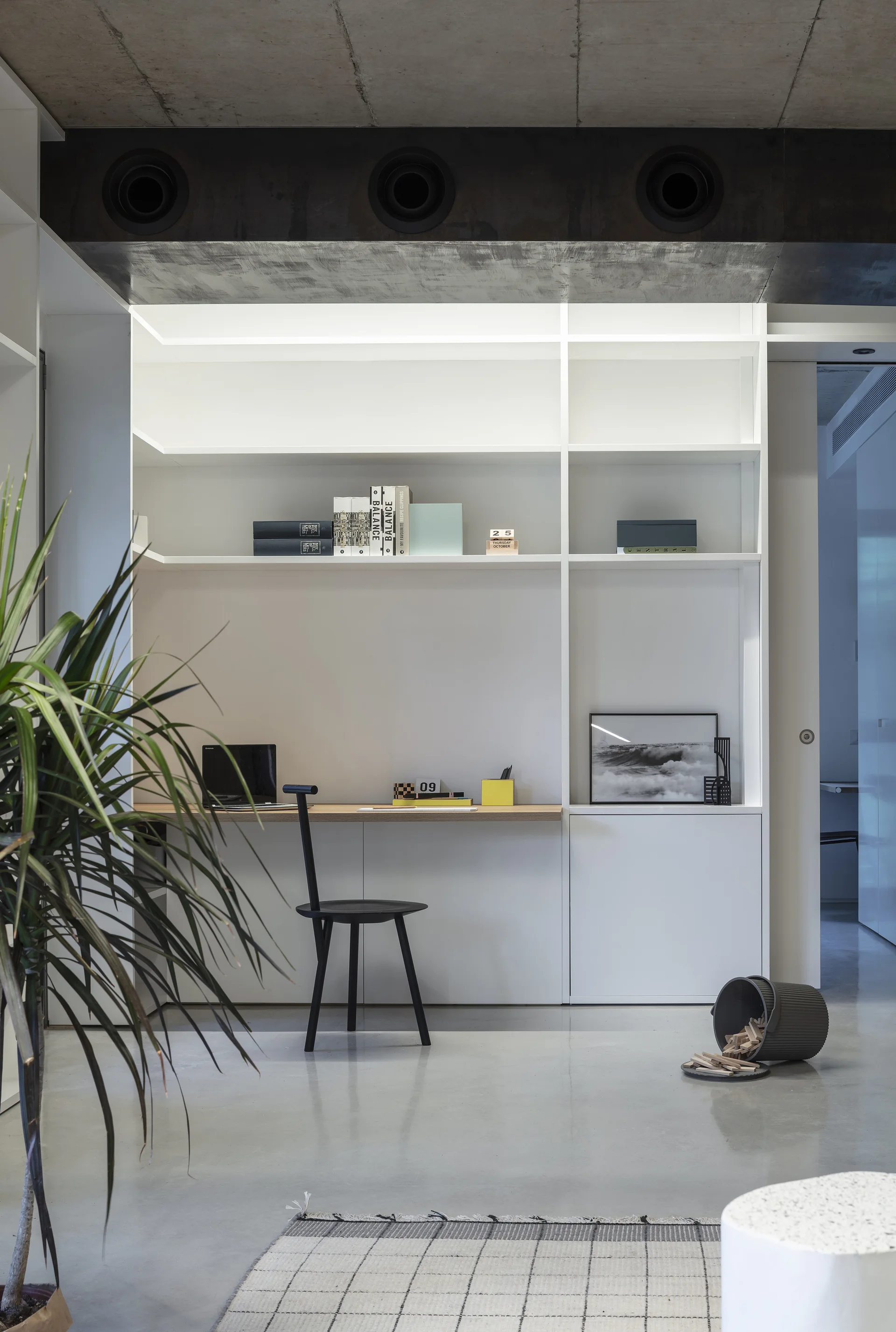 A minimalist home office features a white built-in desk with a wooden top, a black chair, open shelving, and an exposed concrete ceiling.