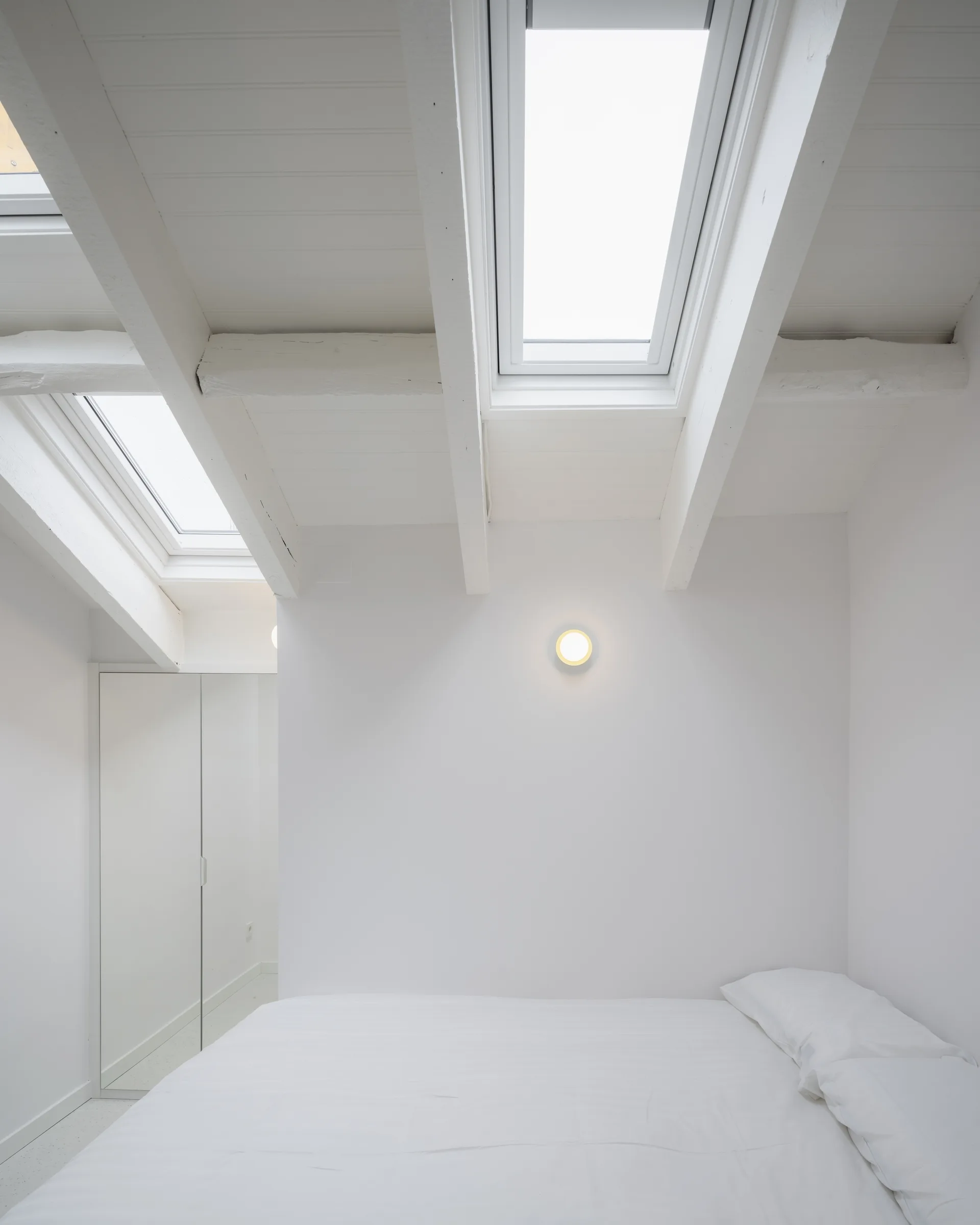 A minimalist white attic bedroom featuring exposed painted ceiling beams, dual skylights, and a clean white bed.