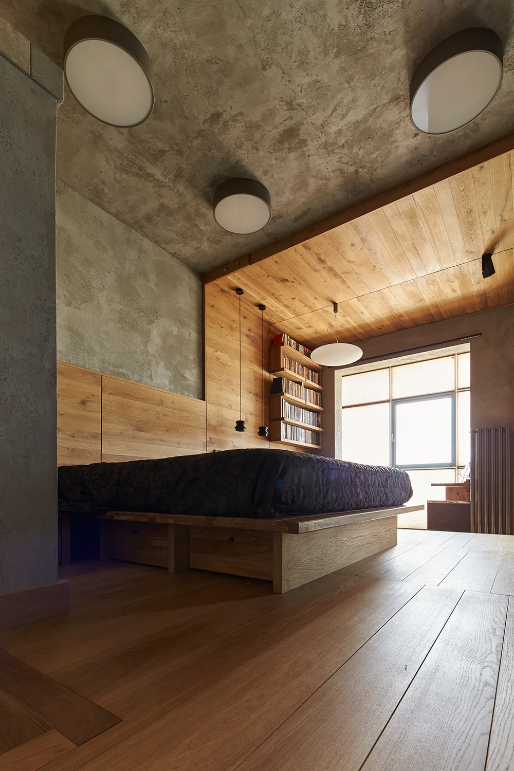 A minimalist bedroom features a wooden platform bed with matching wall panels, a textured concrete ceiling, and a built-in bookshelf.