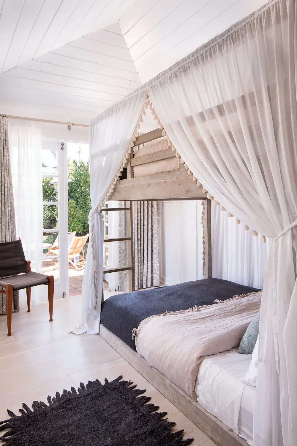 A wooden canopy bunk bed with sheer white curtains sits in a bright room under a vaulted shiplap ceiling.