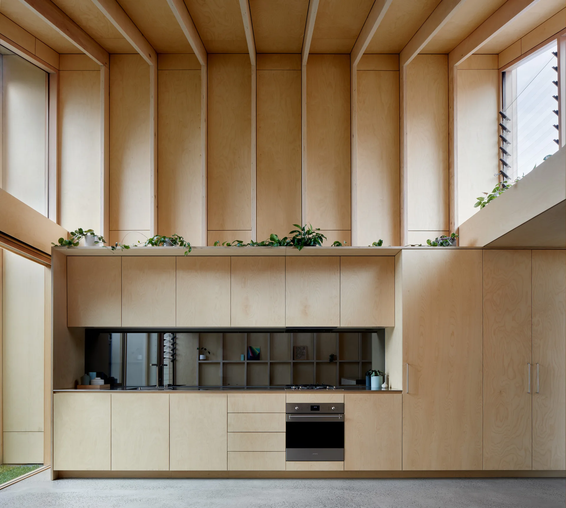 A modern kitchen featuring light plywood cabinetry, a concrete floor, and high ceilings with vertical wooden structural beams.