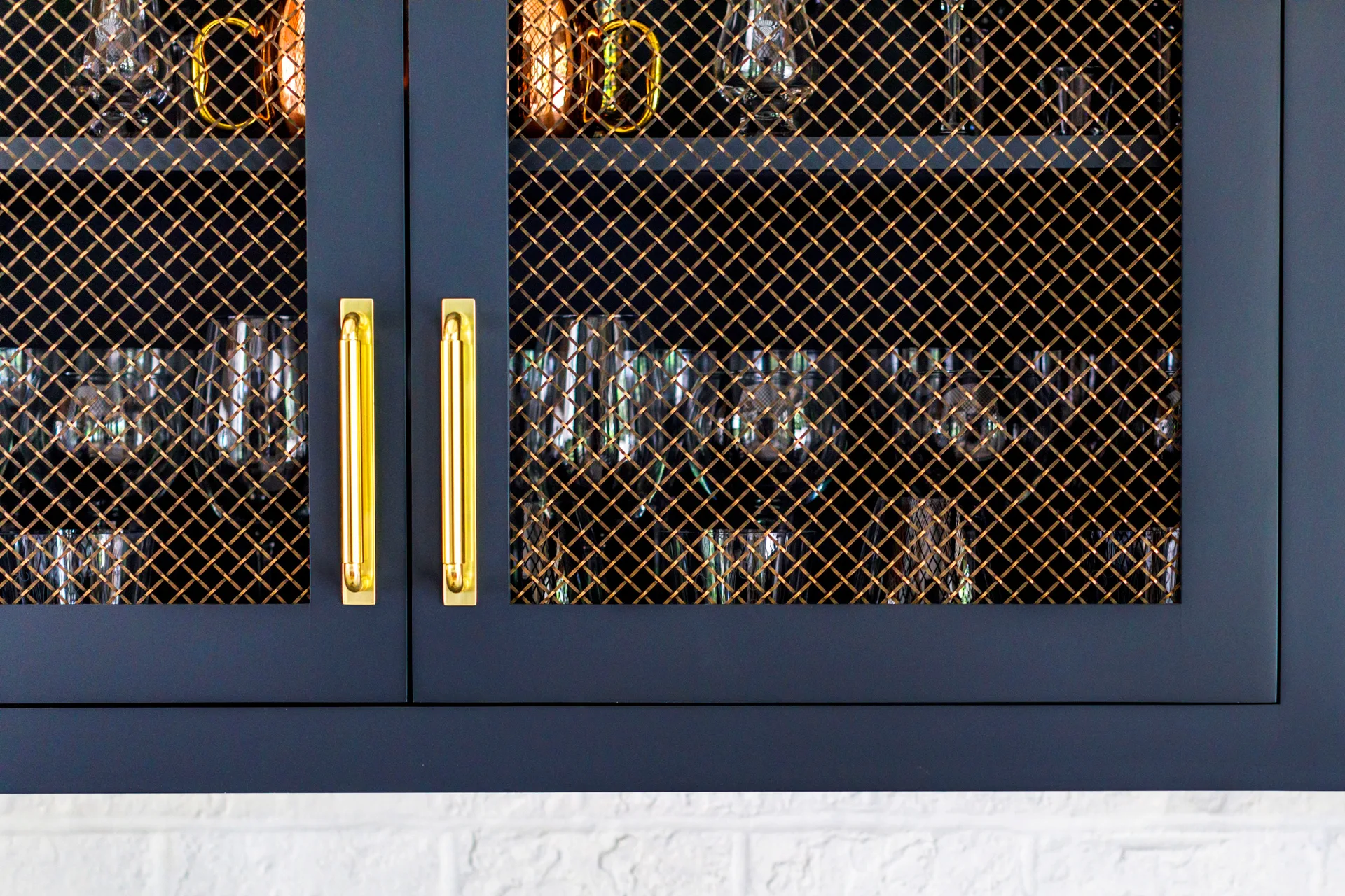 Close-up of navy blue kitchen cabinets featuring gold bar pulls and brass wire mesh door inserts that reveal glassware inside.