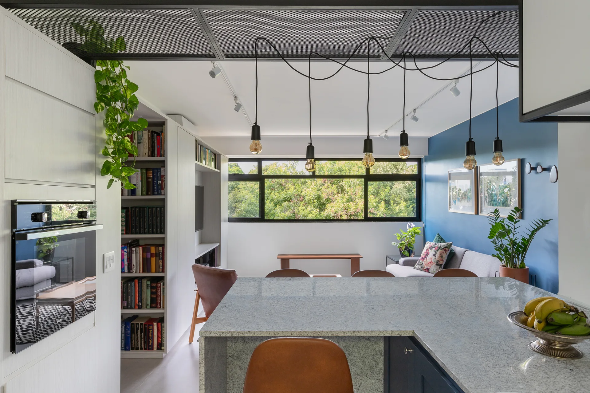 A modern open-plan kitchen featuring a stone-topped island with black pendant lights hanging below an industrial metal ceiling mesh.