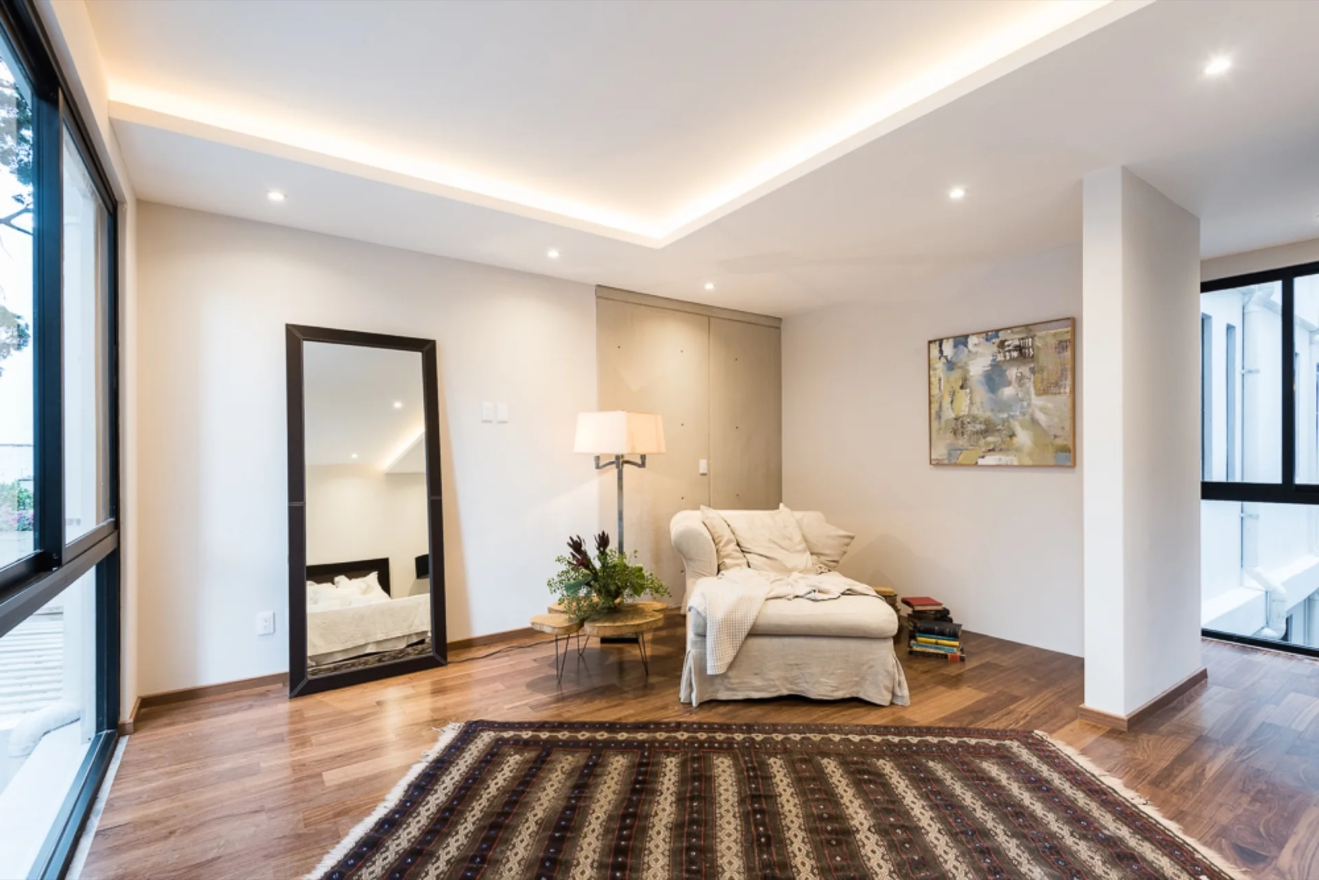 An airy lounge space features an upholstered beige armchair, a large leaning floor mirror, and a geometric patterned rug on hardwood floors.