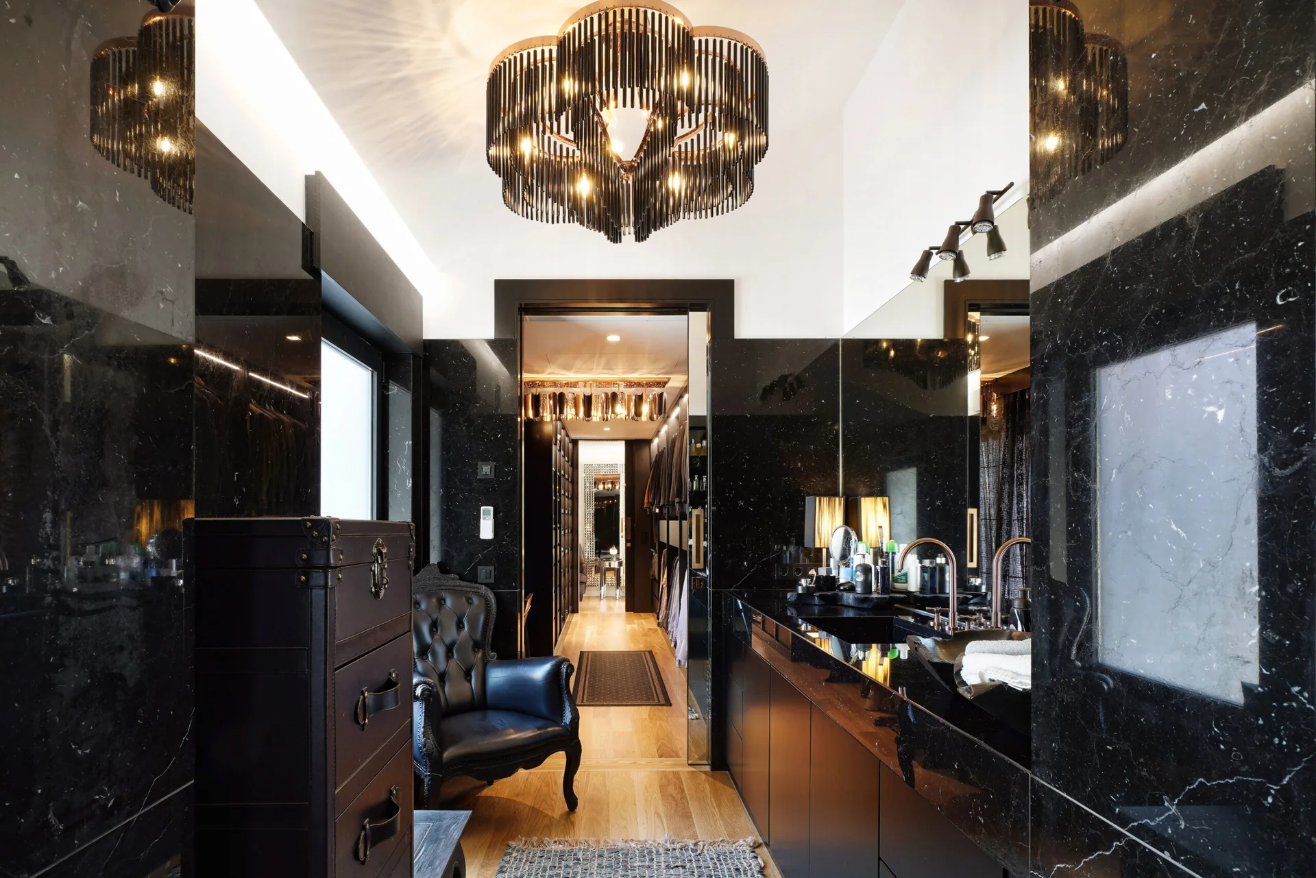 A black marble bathroom features a dark vanity, a black leather armchair, a large tiered chandelier, and a doorway leading into a walk-in closet.