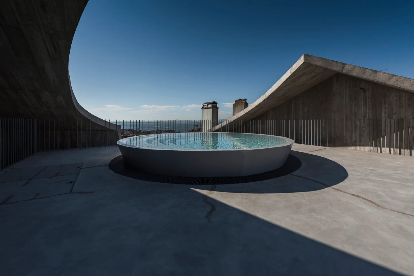 A circular outdoor pool sits on a grey concrete rooftop terrace surrounded by sloped concrete architectural walls under a clear blue sky.