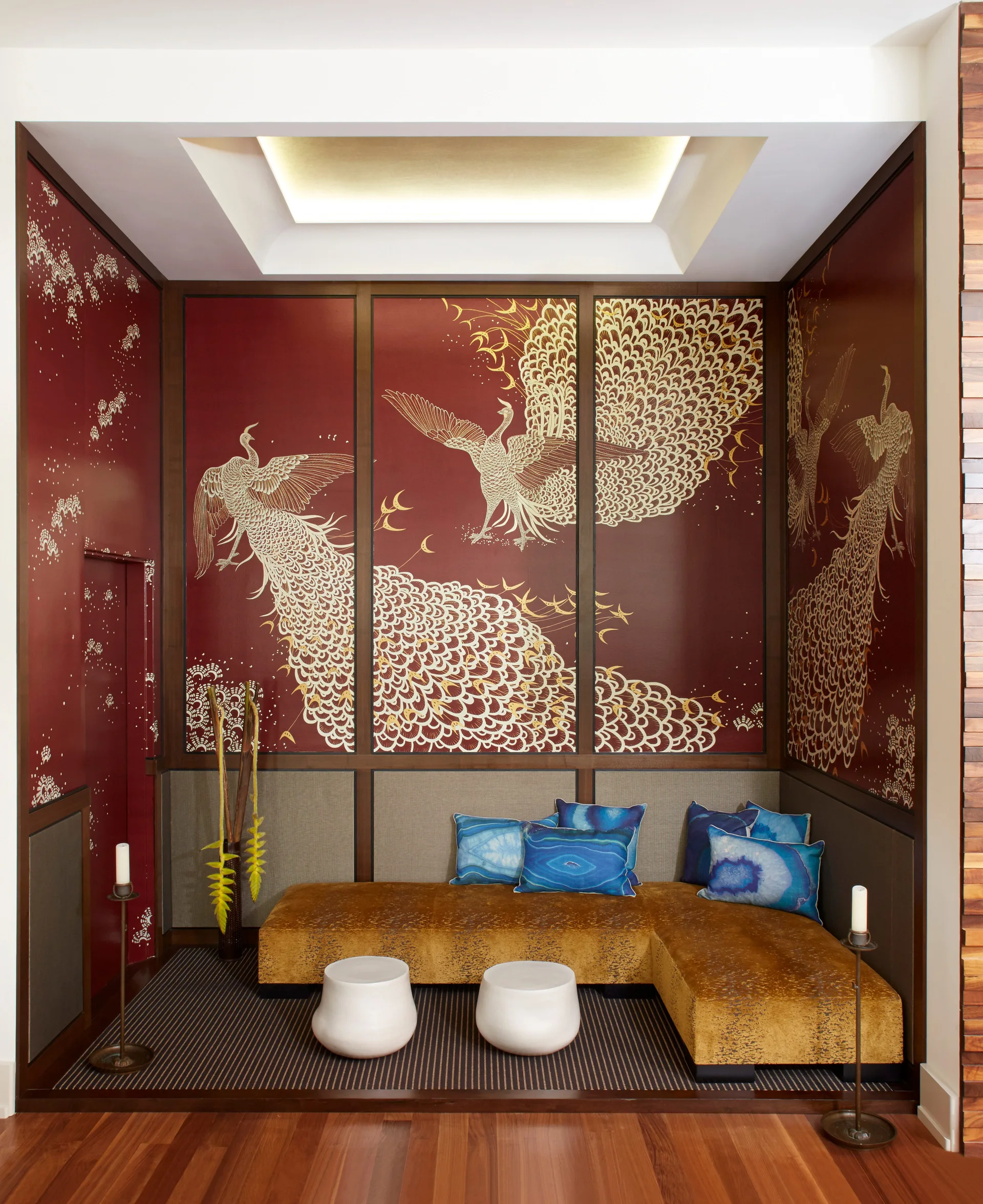 A seating alcove features red wall panels with gold peacock illustrations behind a low-profile yellow sofa, blue throw pillows, and two white stools.
