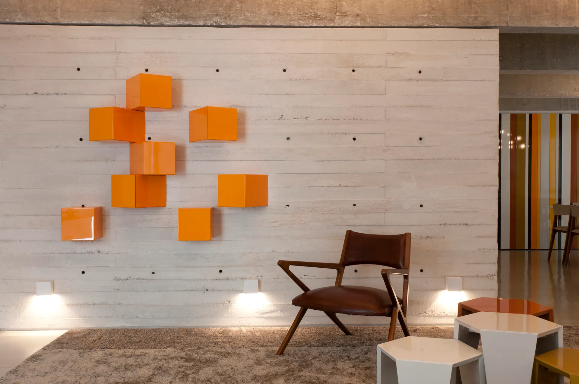 A mid-century brown leather armchair sits in front of a textured concrete wall decorated with bright orange cube shelves.