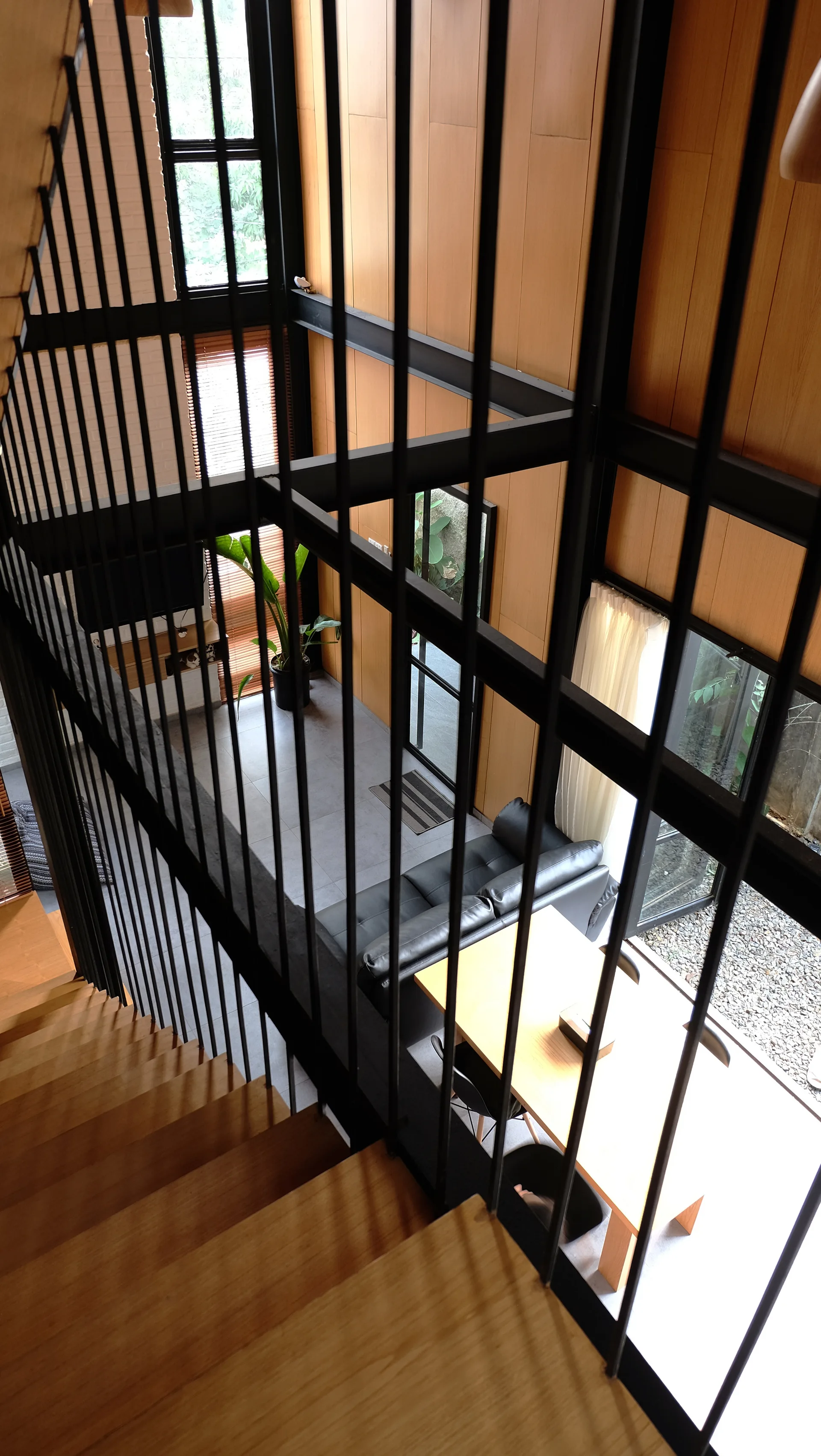 A high-angle view from a wood staircase looking down at a modern living area with vertical wood paneling, a black leather sofa, and a dining table.