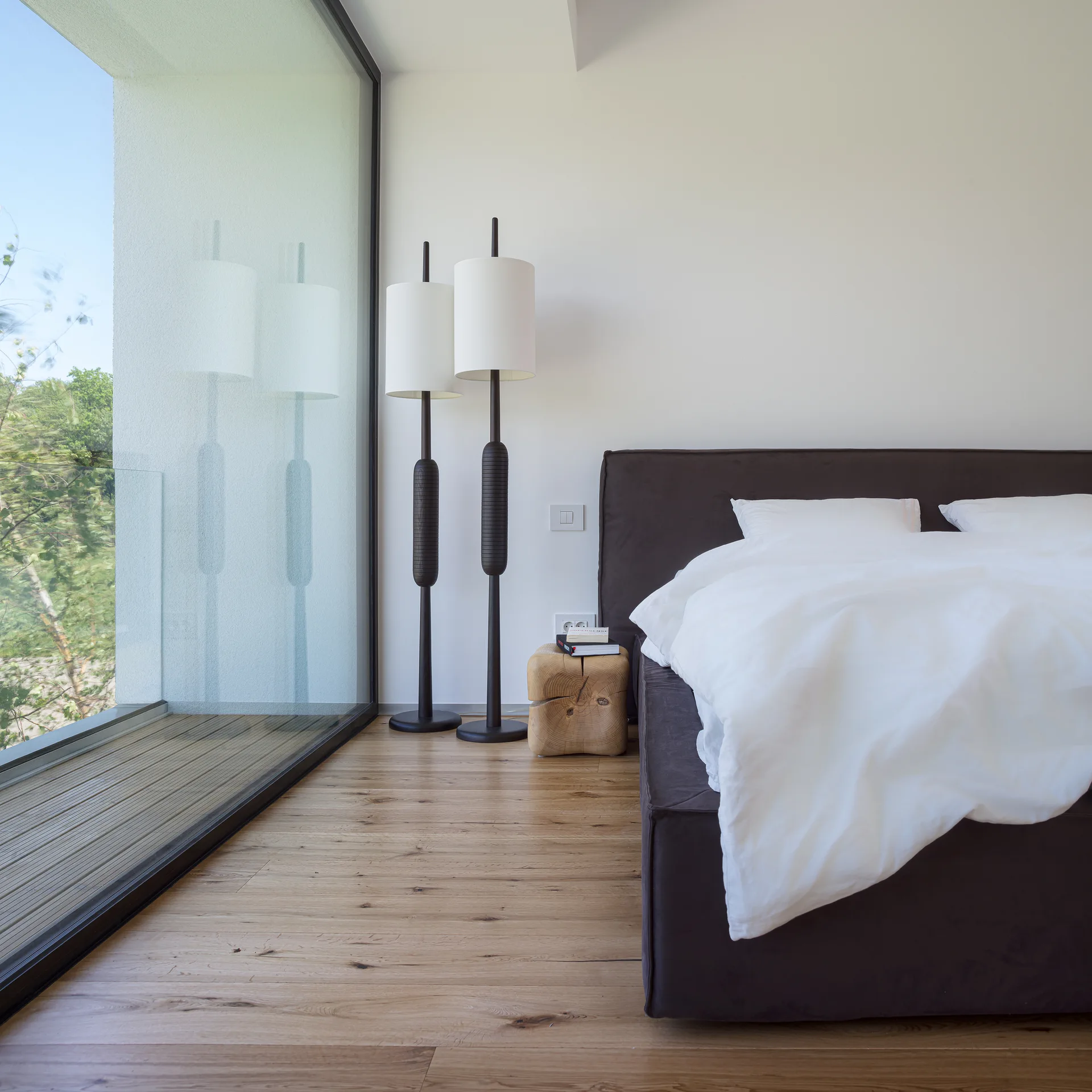 A modern bedroom features a dark upholstered bed next to two tall black floor lamps, a wood block nightstand, and a floor-to-ceiling glass door.