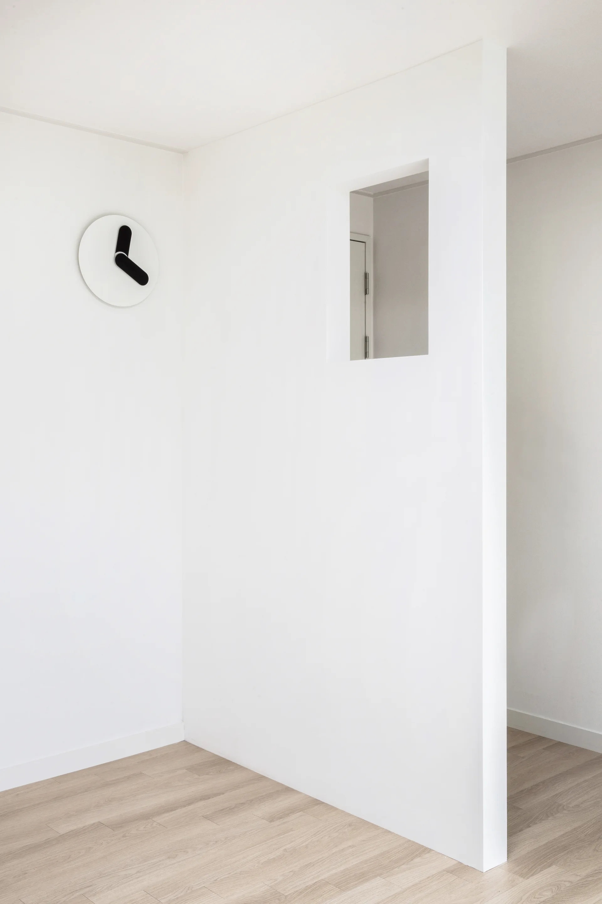A minimalist white partition wall with a square cutout window stands on light wood floors next to a simple white wall clock.