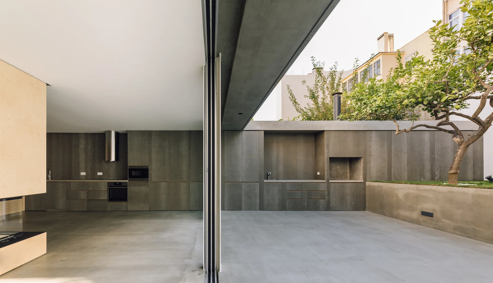 A modern indoor-outdoor kitchen featuring seamless grey concrete wall panels, flat cabinet doors, and a large sliding glass partition.