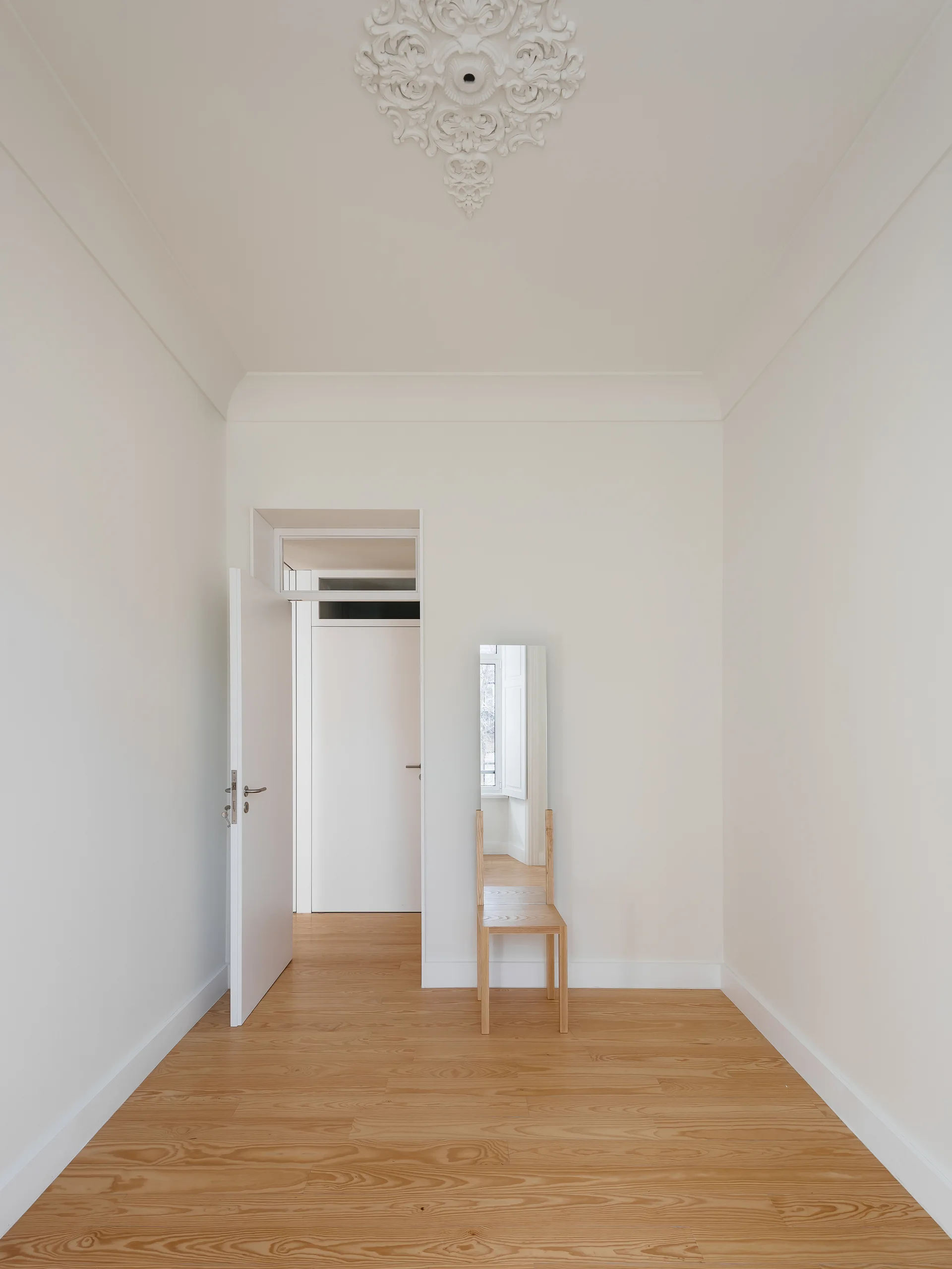 A minimalist white hallway with light wood flooring features an open white door and a single wooden chair beneath a tall thin mirror.