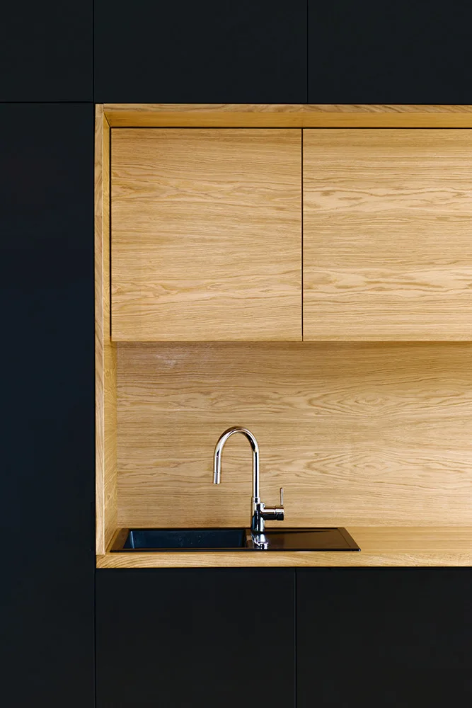 A modern kitchen corner featuring matte black cabinets surrounding a recessed natural wood niche with a black sink and chrome arched faucet.