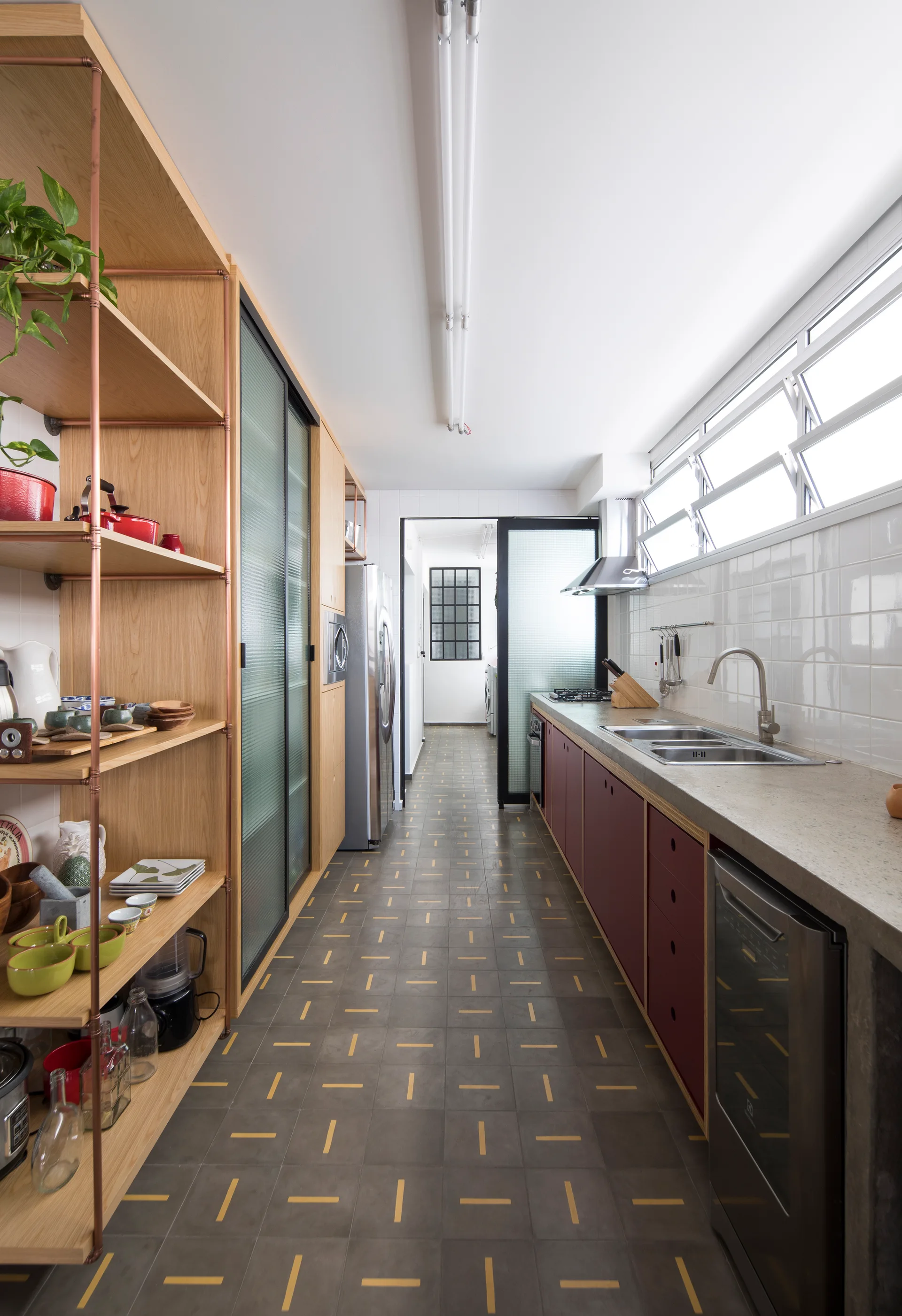 A narrow galley kitchen features red base cabinets, a concrete countertop with a double sink, and wooden shelves supported by copper piping.