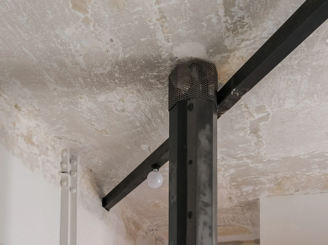A raw steel structural column and black horizontal beam meet an aged, distressed plaster ceiling with a small exposed white globe bulb.