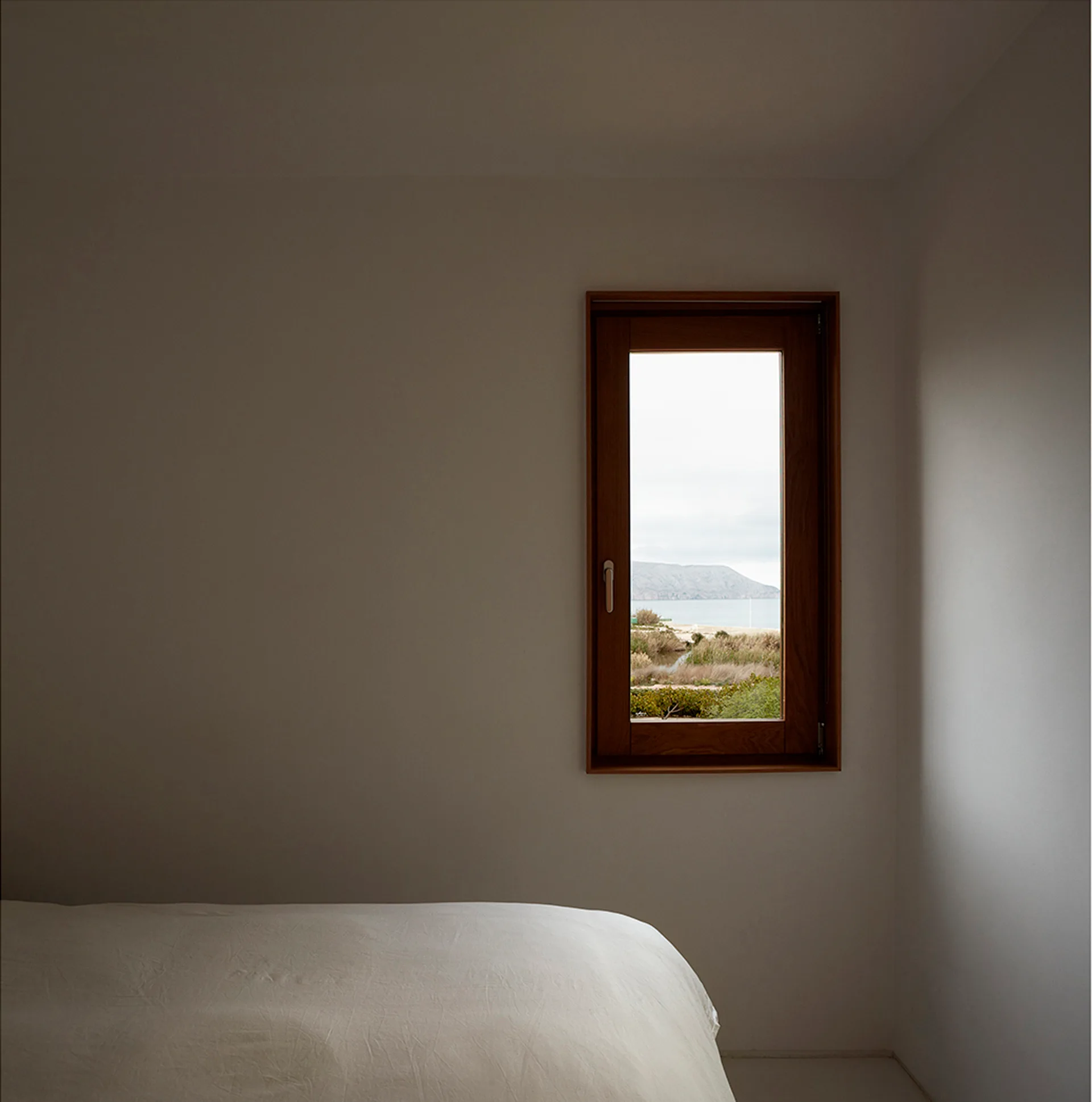 A minimalist white bedroom features a single wood-framed window showing a coastal landscape view next to a bed with white sheets.