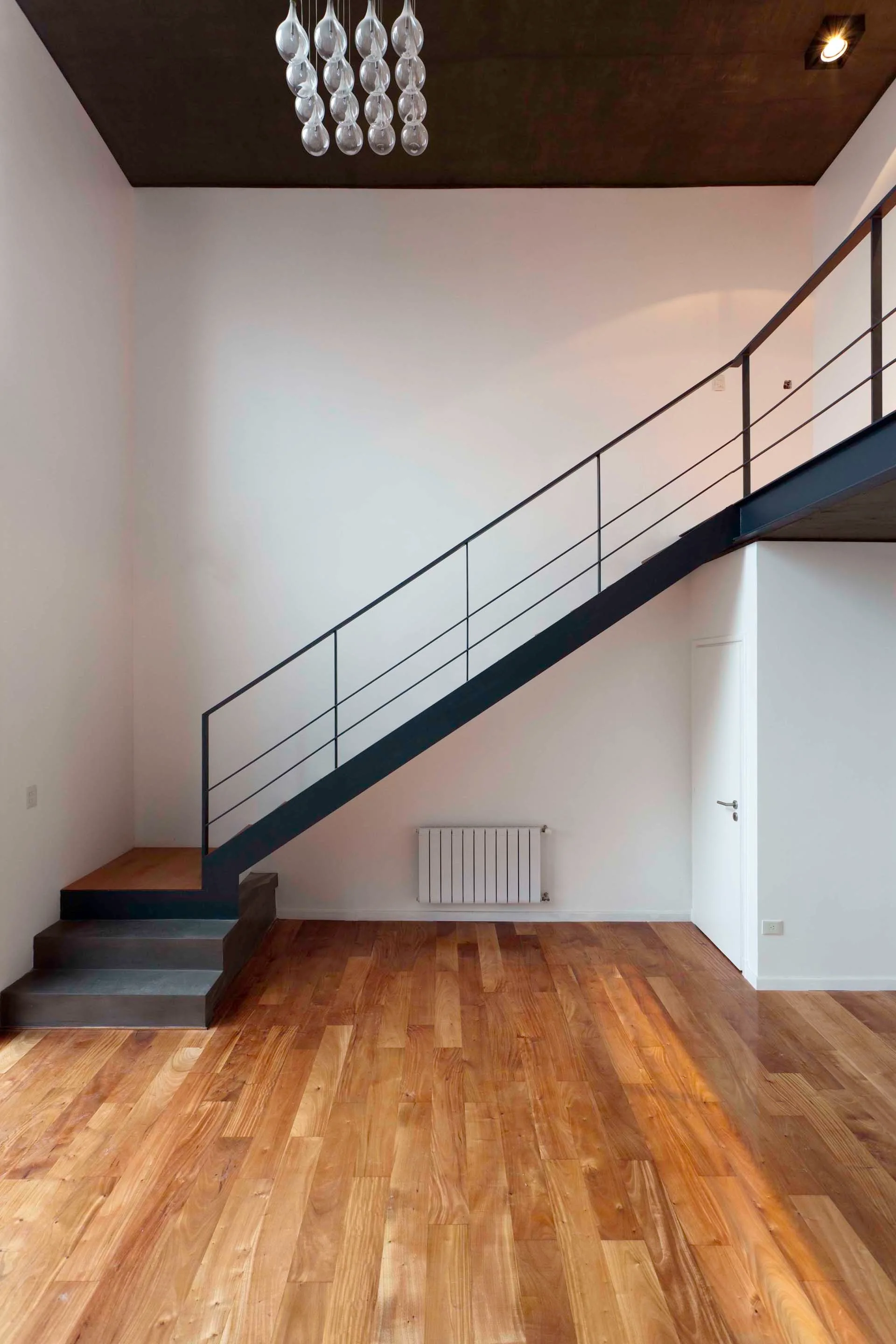 A black metal staircase with wood treads leads to a mezzanine level in an airy room with white walls and warm wood plank flooring.