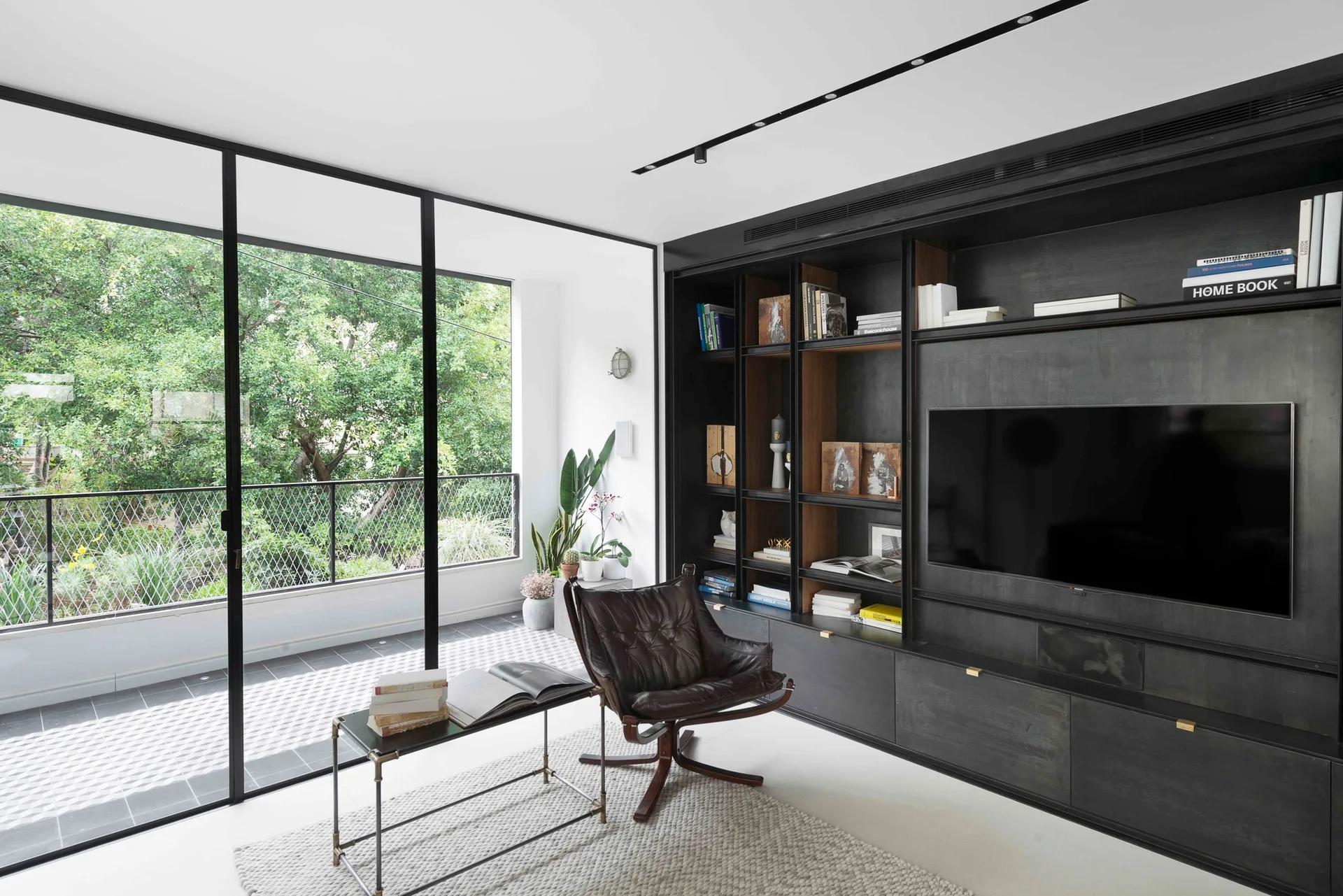 A modern living room features a dark built-in media unit, a brown leather lounge chair on a textured rug, and floor-to-ceiling glass doors.