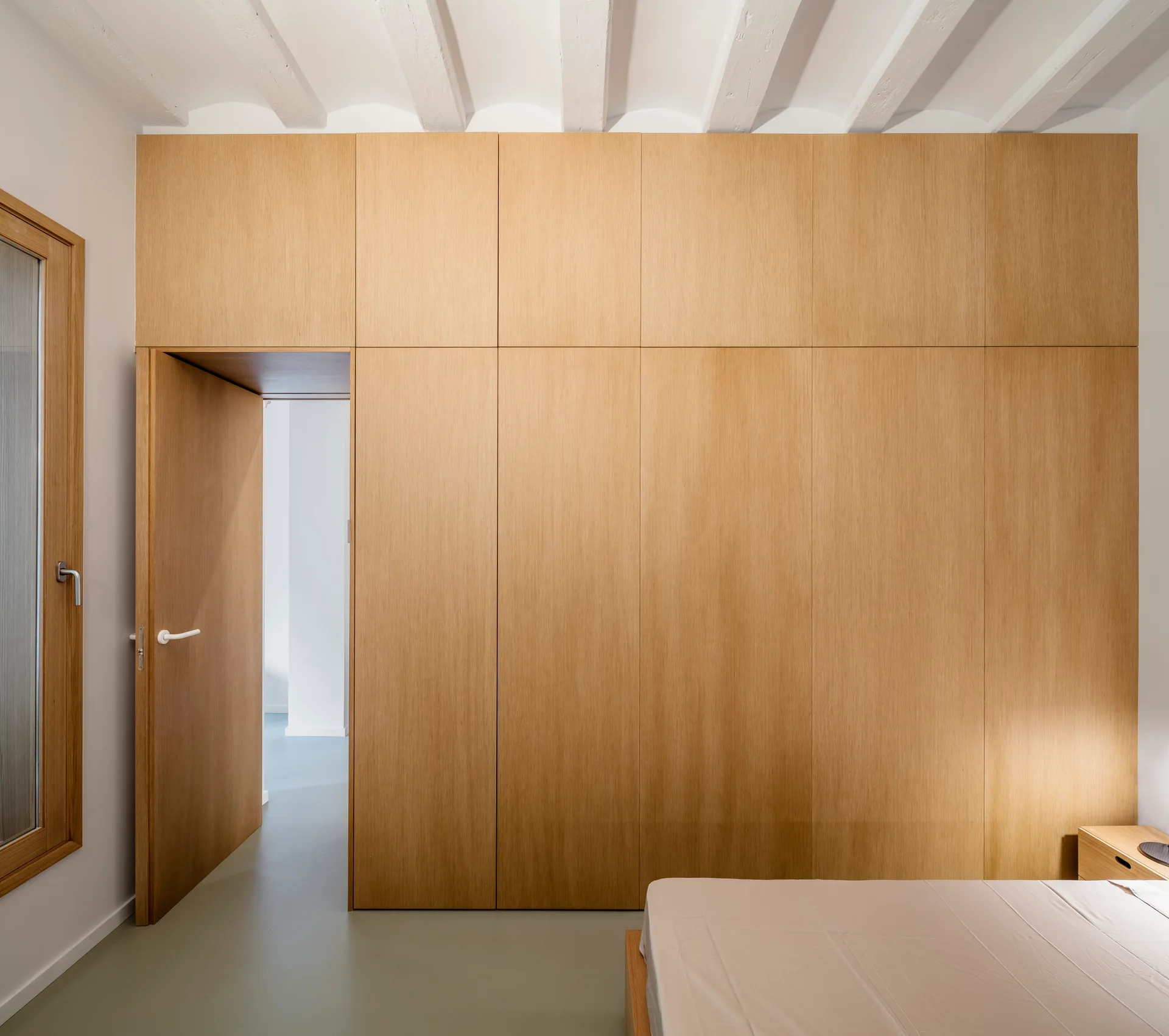 A minimalist bedroom featuring a floor-to-ceiling light wood built-in wardrobe wall with an integrated door, white ceiling beams, and a simple bed.