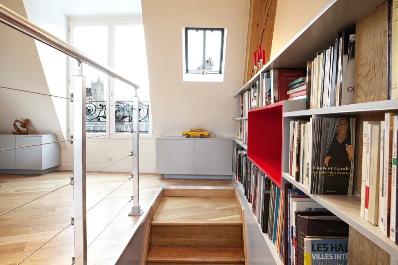 An attic landing features wood stairs with a metal railing, built-in bookshelves with a red insert, and natural light from a window and skylight.