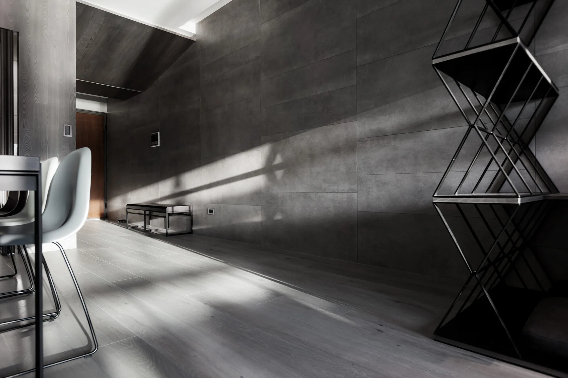 A modern minimalist corridor with large dark grey stone wall panels, a black geometric metal shelving unit, and sunlight casting sharp diagonal shadows.