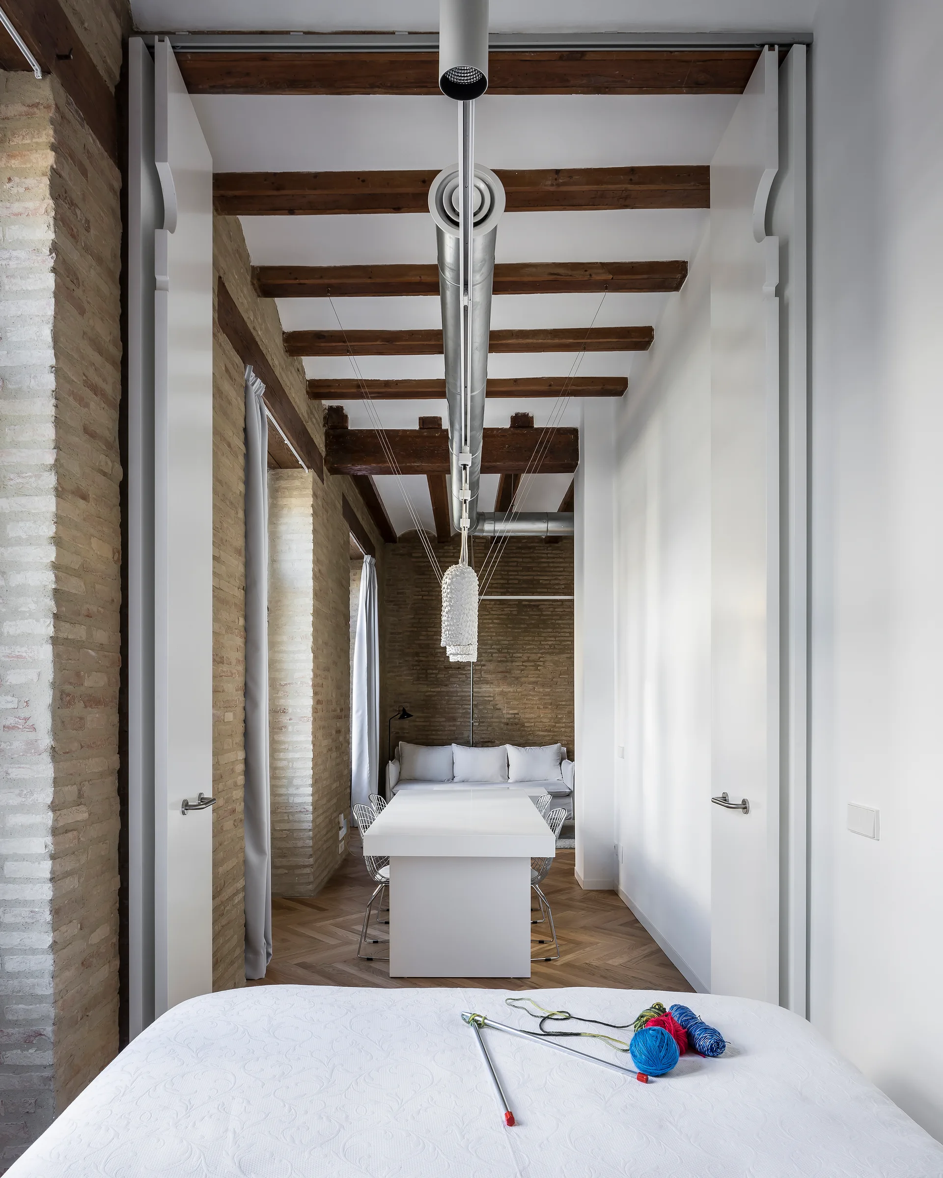 A bedroom view looking toward a white dining table in a loft featuring exposed brick walls, timber ceiling beams, and sliding white door panels.