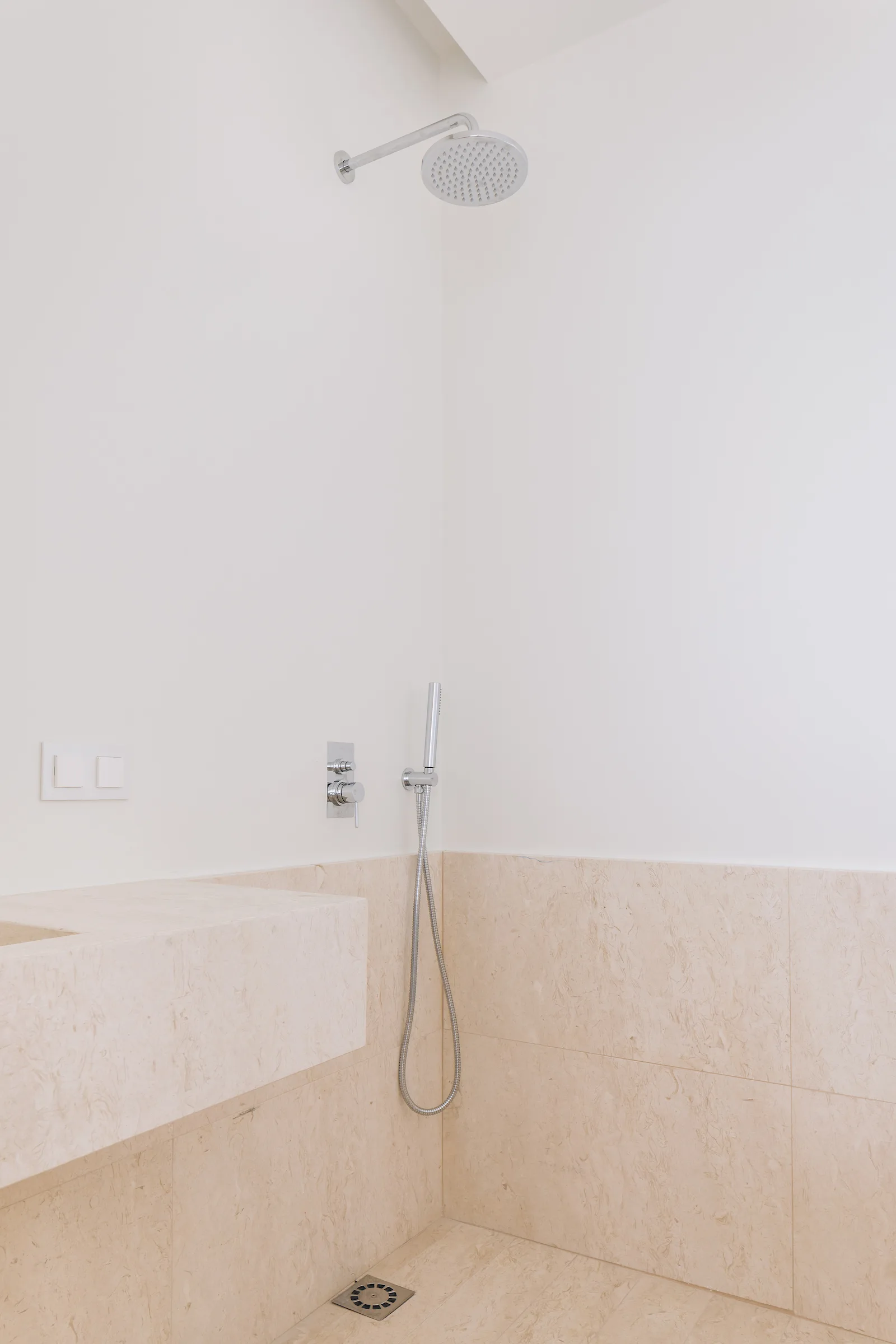 A minimalist walk-in shower featuring beige stone tile on the lower wall and floor, white upper walls, and chrome fixtures.