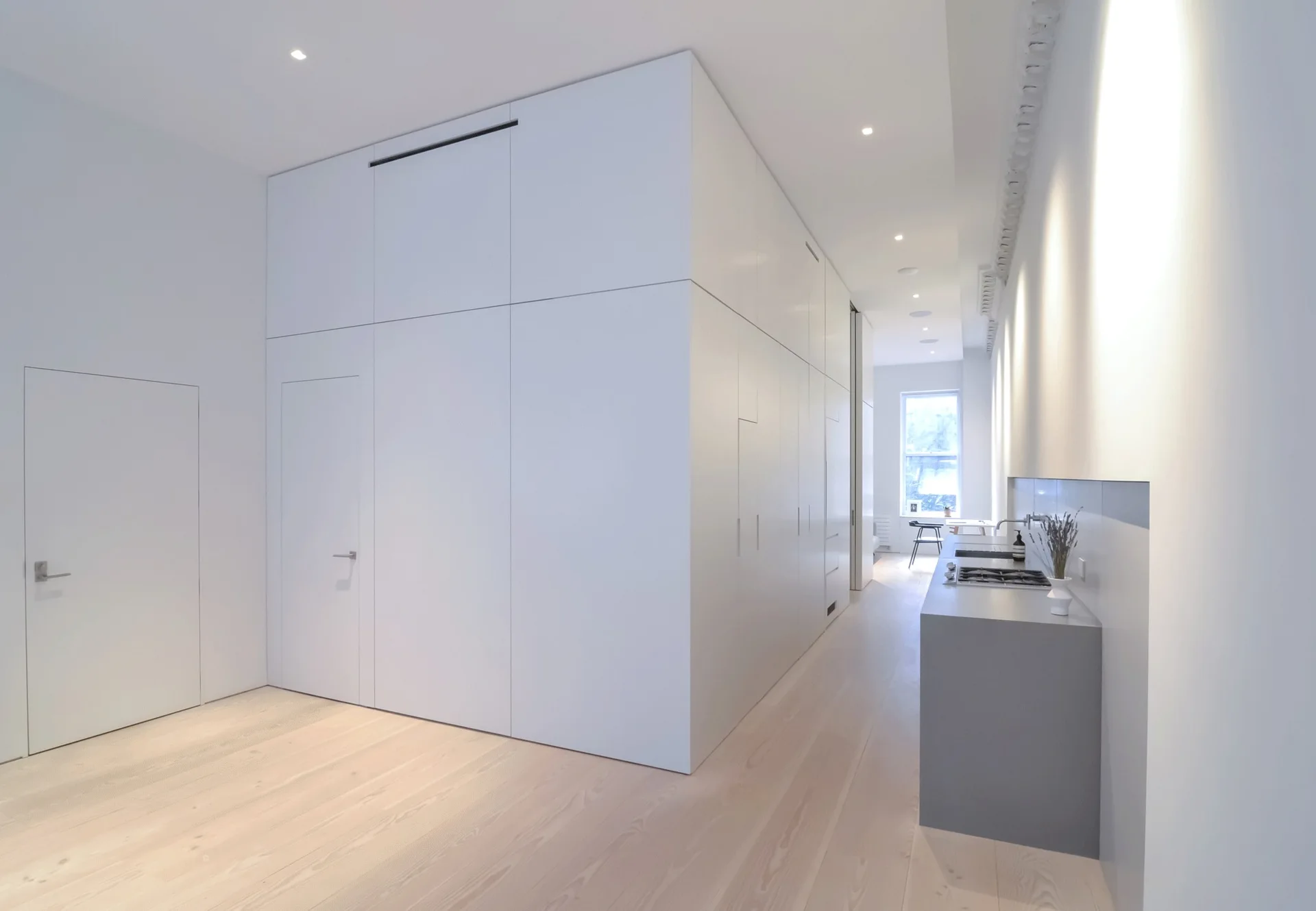 A minimalist kitchen features a massive white wall of integrated cabinetry, a grey kitchen island, and light wood flooring.