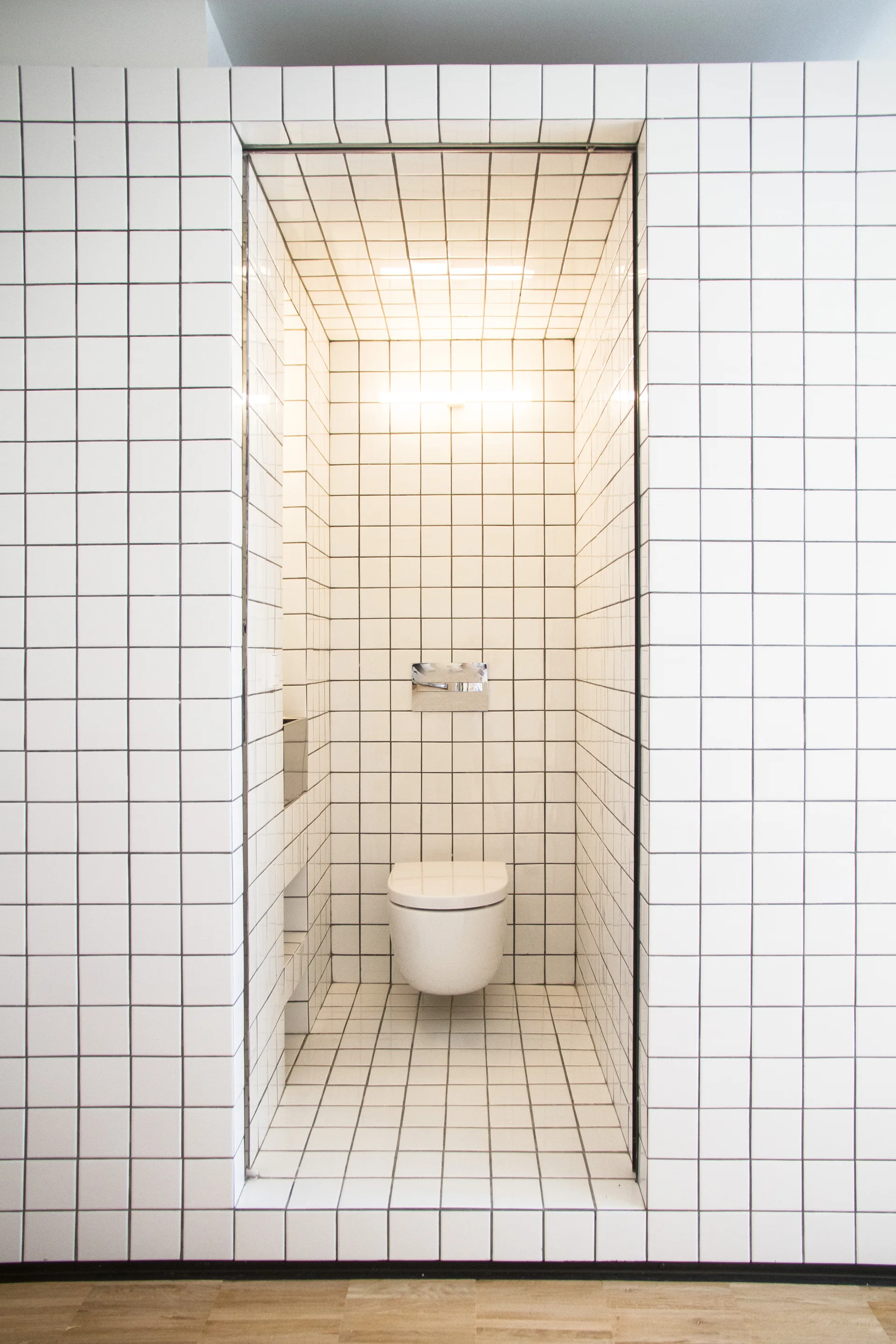 A minimalist powder room features a wall-mounted toilet inside a recessed alcove covered entirely in white square grid tiles with dark grout.