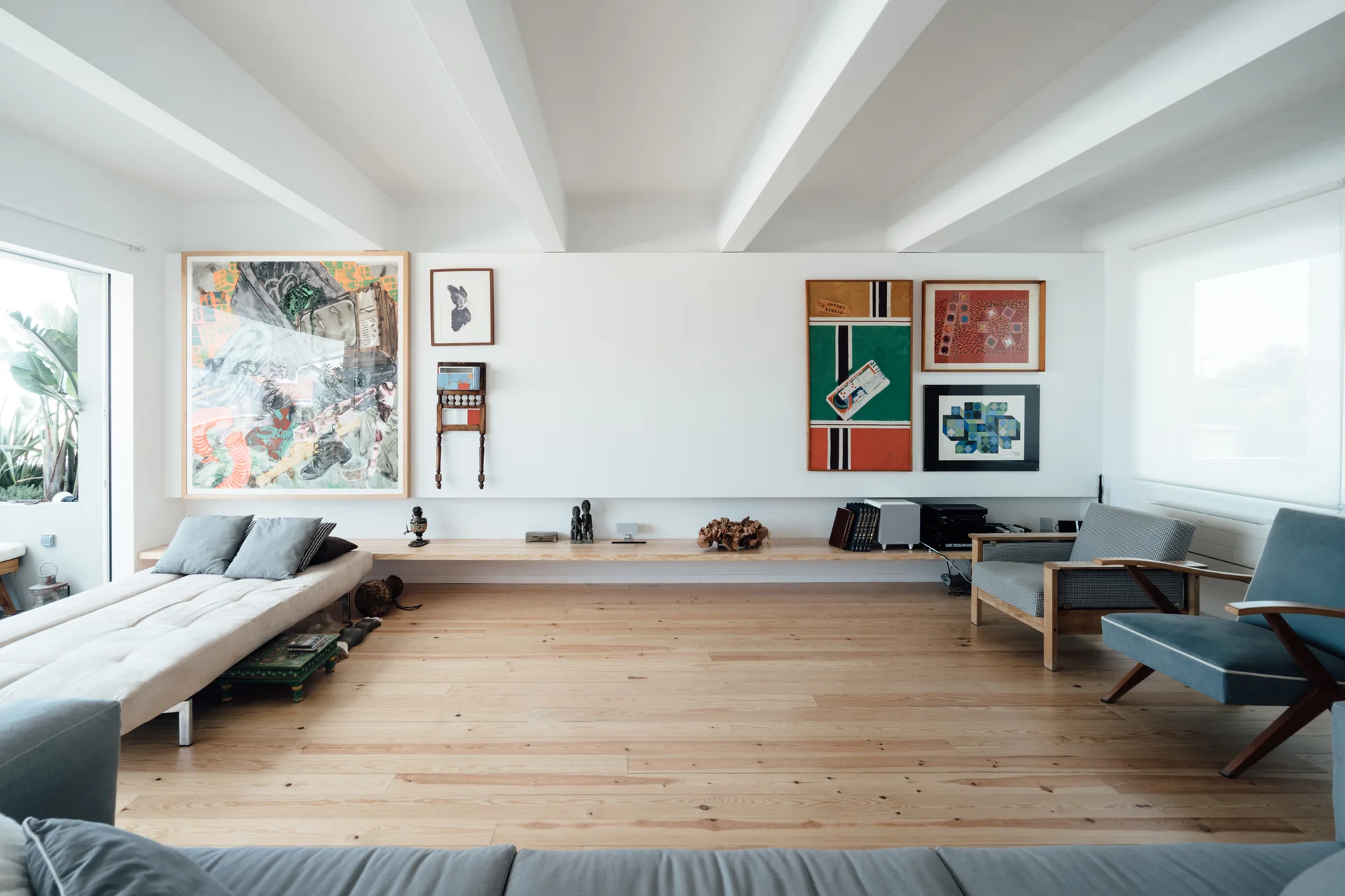 A minimalist white living room features a long light wood floating shelf, framed artwork on the walls, and prominent white ceiling beams over wood floors.