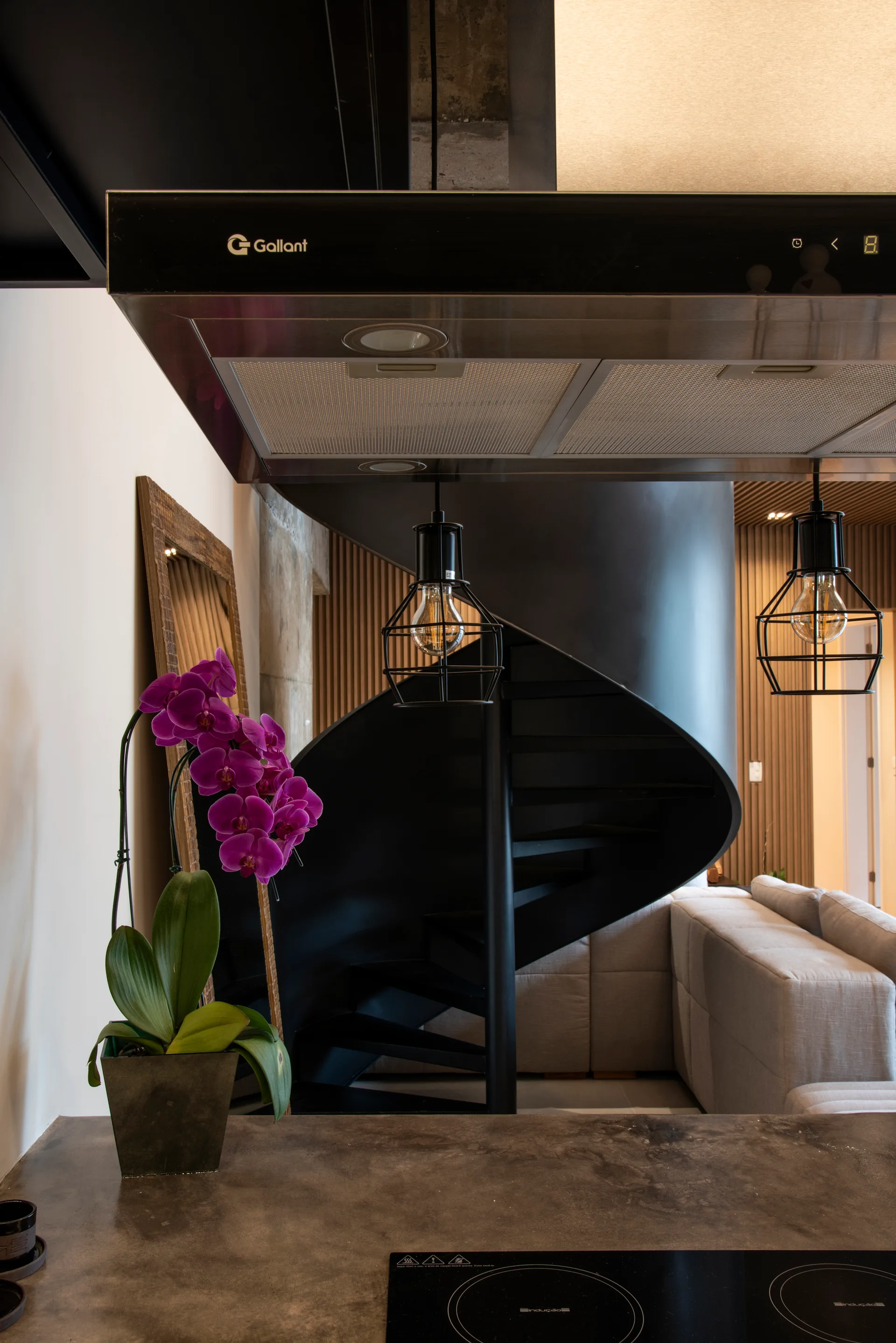 A dark kitchen counter with a purple orchid overlooks a black metal spiral staircase and a light grey sofa in a modern open home.