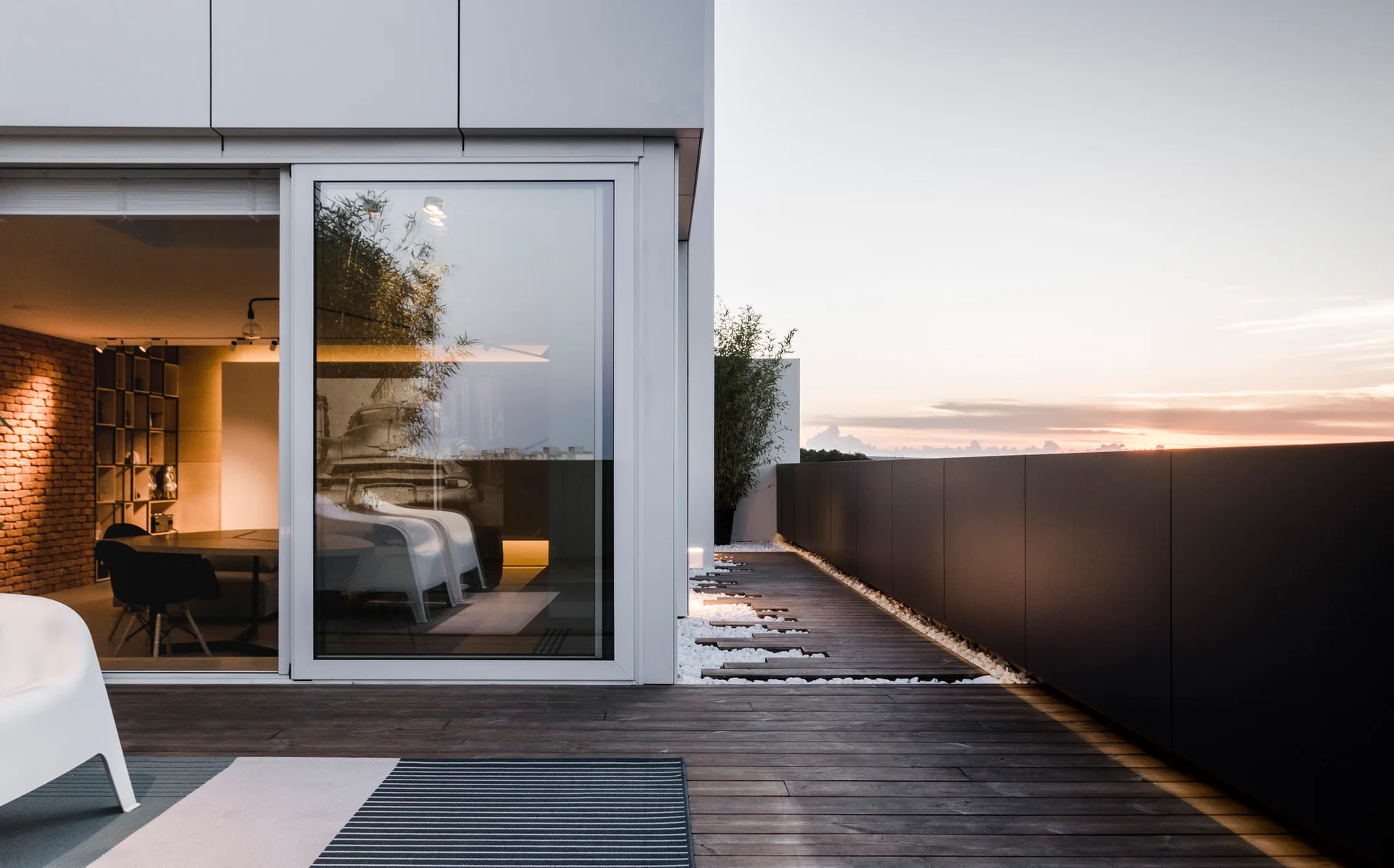A modern outdoor terrace features dark wood decking, a solid gray railing, and a large sliding glass door revealing a lit indoor room.