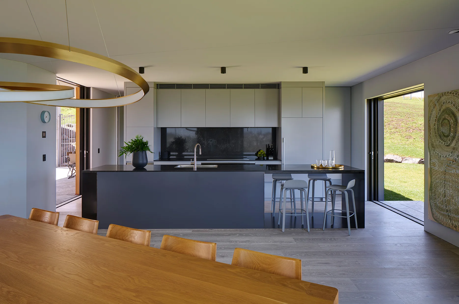 A minimalist kitchen features a dark gray island with stools, flat-front gray cabinetry, and a light wood dining table in the foreground.